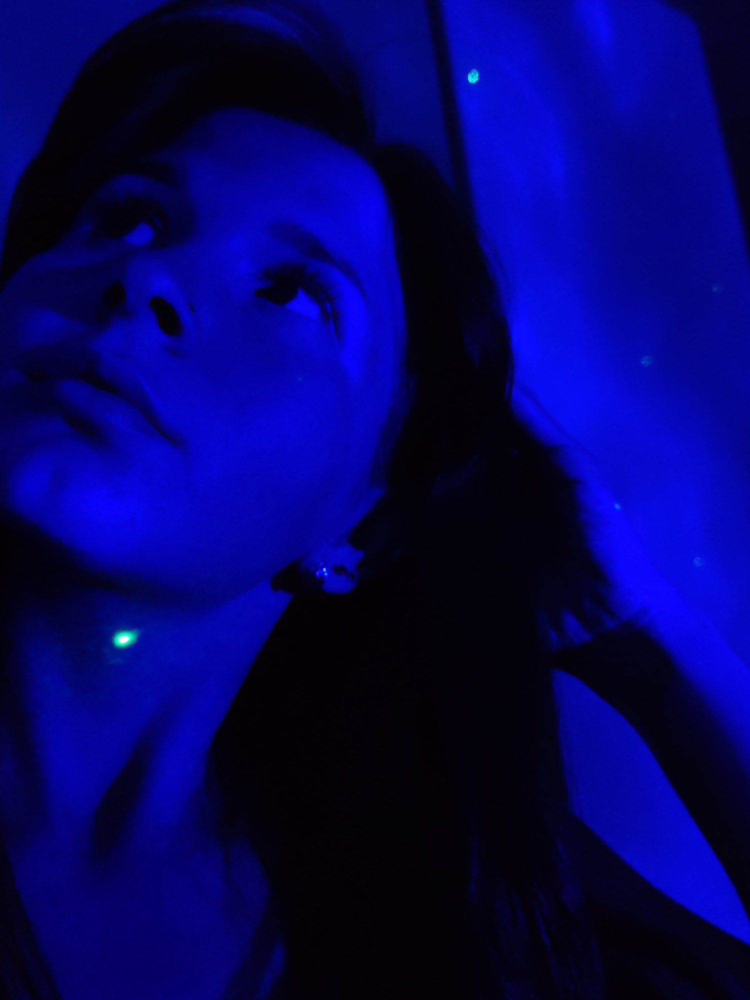 A person is shown in a low-angle view, illuminated by a strong blue light. The person's face is visible, looking upward. A green dot of light is visible on the neck. The background is dark and indistinct, with a few small points of light suggesting a room or space. There is no indication of the location being in Фоки, Russia.