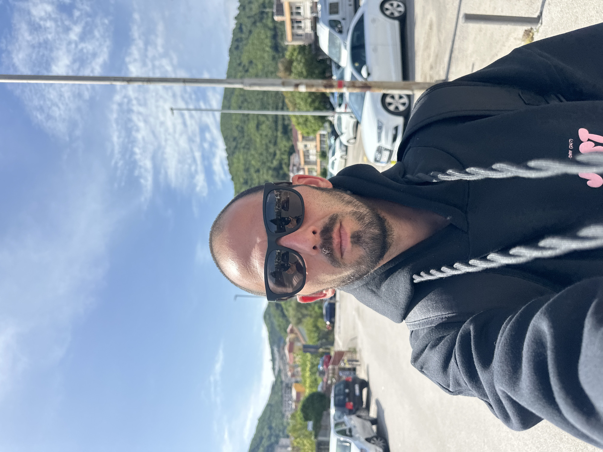 The image is a rotated selfie of a man with a shaved head and beard, wearing sunglasses and a black hooded sweatshirt. He is standing outdoors, presumably in Budva, Montenegro, given the presence of a building with terracotta roofs and a green, mountainous backdrop typical of the region. Multiple white cars are visible, parked along a sidewalk next to the road. The sky is blue with scattered white clouds. A metal pole stands in the background. The image has been rotated 90 degrees counter-clockwise.