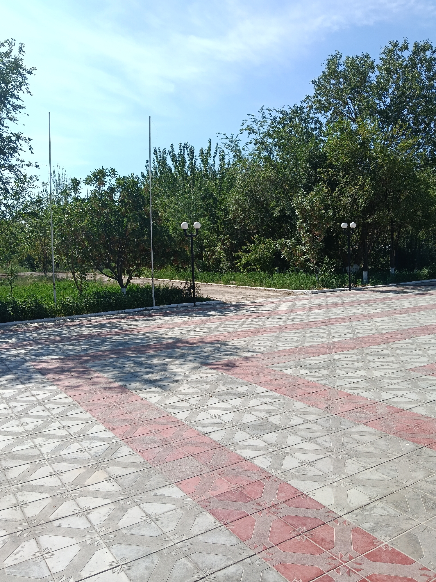 A paved plaza with red and white patterned tiles stretches towards a line of trees under a bright blue sky with sparse clouds. Two tall, slender poles stand vertically against the sky to the left of center, while two white globe-shaped lamps on a black pole are visible among the trees to the right. Shadows from unseen foliage dapple the patterned tiles. The location is identified as Kungrad, Uzbekistan.