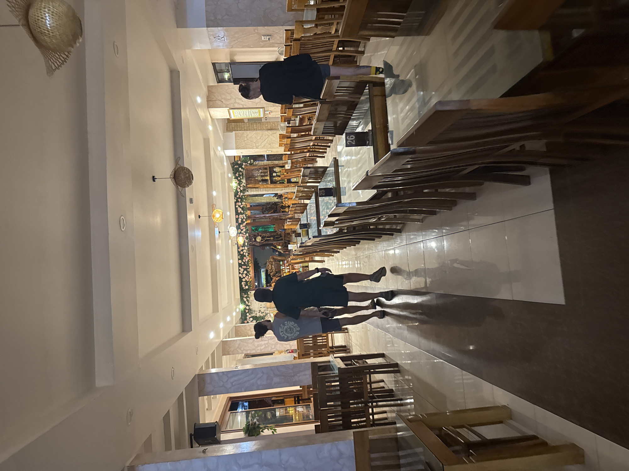 The photograph depicts the interior of a restaurant in Tú Lệ, Vietnam. The space is characterized by rows of wooden tables and chairs extending into the distance. Two individuals are walking down the aisle between the tables. The decor includes hanging lamps and floral arrangements toward the far end of the restaurant. The flooring is tiled with a contrasting pattern.