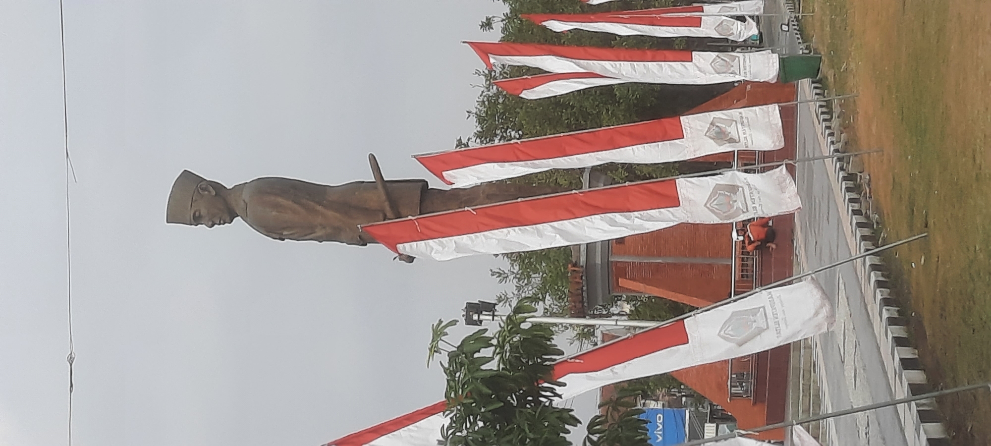The image shows a statue of a man in a uniform and cap, holding a sword. The statue is large and stands tall against an overcast sky. In front of the statue, there are several red and white flags, which appear to be the Indonesian flag. The flags are arranged in a line and are fluttering in the wind. In the background, there is a brick structure and some trees. A person in an orange shirt and white helmet is sitting on a ledge in front of the brick structure. The scene is likely a public space in Kanigoro, Indonesia, possibly a monument or memorial.