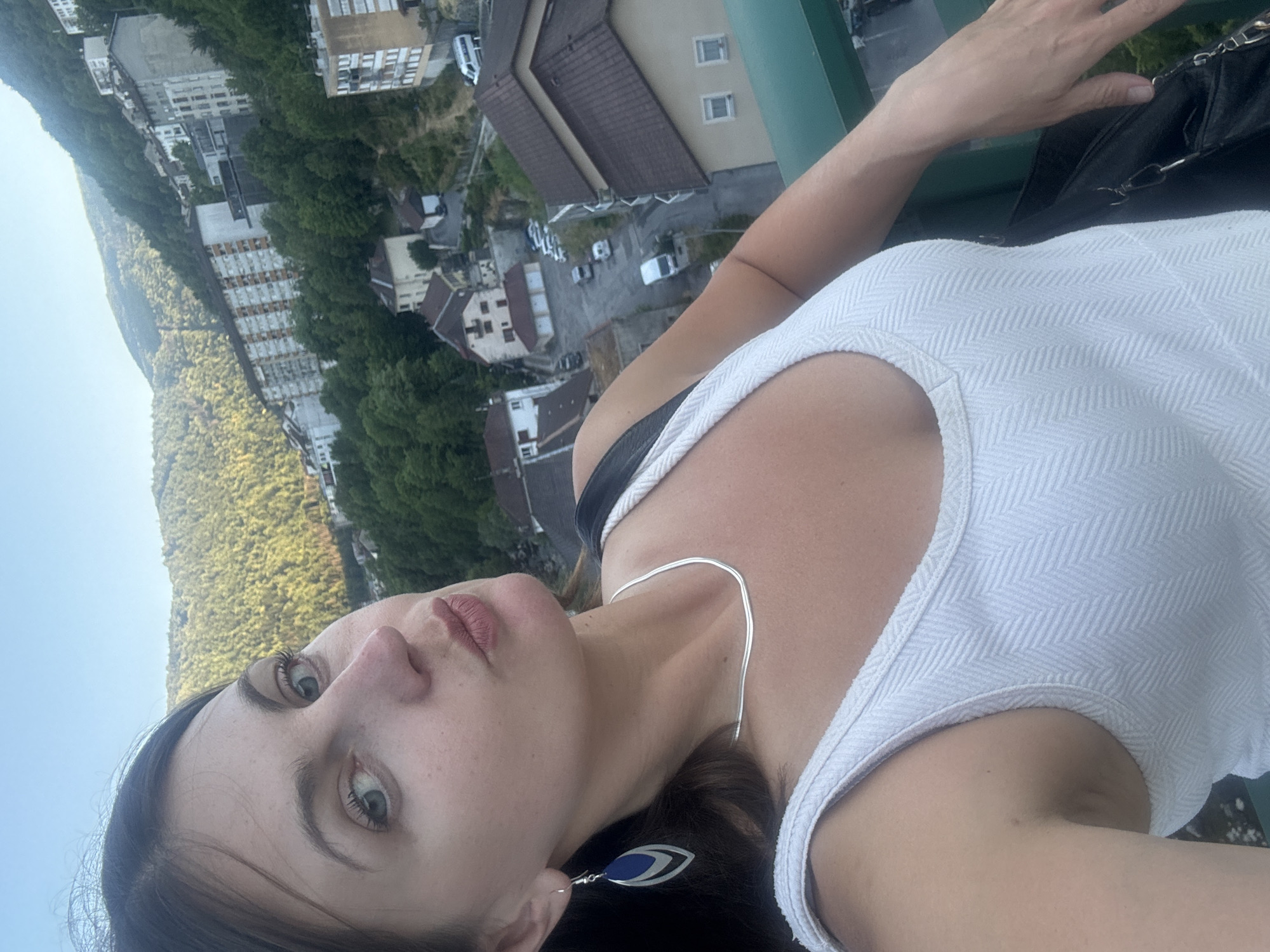 A woman is taking a selfie outdoors, her face taking up the left half of the frame, with her head tilted and lips pursed. She has long, dark brown hair, blue eyes, and is wearing a white tank top, a silver necklace, and blue and silver earrings. Behind her, a hillside town is visible, characterized by buildings of various heights, some with dark tiled roofs, nestled amongst lush green trees. The sky is a clear, pale blue. The woman's right arm is visible, resting on a green railing, with a black bag to her side. The scene appears to be a scenic overlook in the city of Saint-Claude, France.