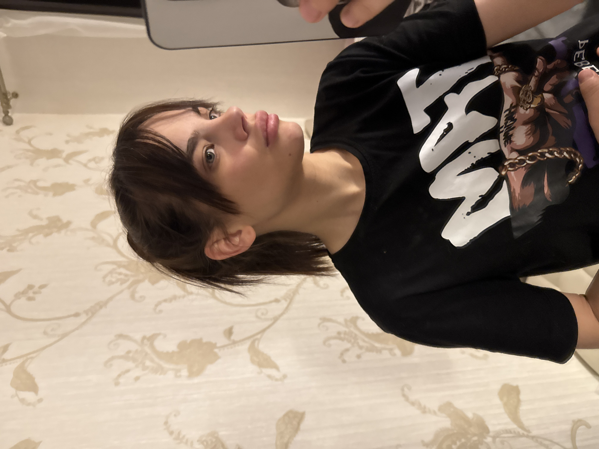 A young person is depicted in a rotated close-up portrait, likely a selfie or mirror shot, taken indoors in Rostov-on-Don, Russia. The individual has fair skin, light-colored eyes, and dark brown hair, which is styled with bangs and pulled back from their face. Their expression is neutral to slightly contemplative, with their gaze directed upwards and to the side, and lips gently parted.
They are wearing a black short-sleeved T-shirt that features a prominent graphic. The left side of the graphic displays large white text, where the letters "AR" are clearly visible, possibly part of the word "ARMY." To the right of this text, there is an illustration of a muscular arm or hand, adorned with several gold rings on the fingers and a thick gold chain wrapped around the wrist. This illustration is set against a purple background section on the shirt.
The background of the shot reveals a light-colored wall decorated with a subtle, ornate floral or vine pattern in a muted beige or gold tone. The lighting appears even, characteristic of an indoor setting. A hand holding a device, possibly a smartphone used to capture the image, is partially visible at the top right of the frame.