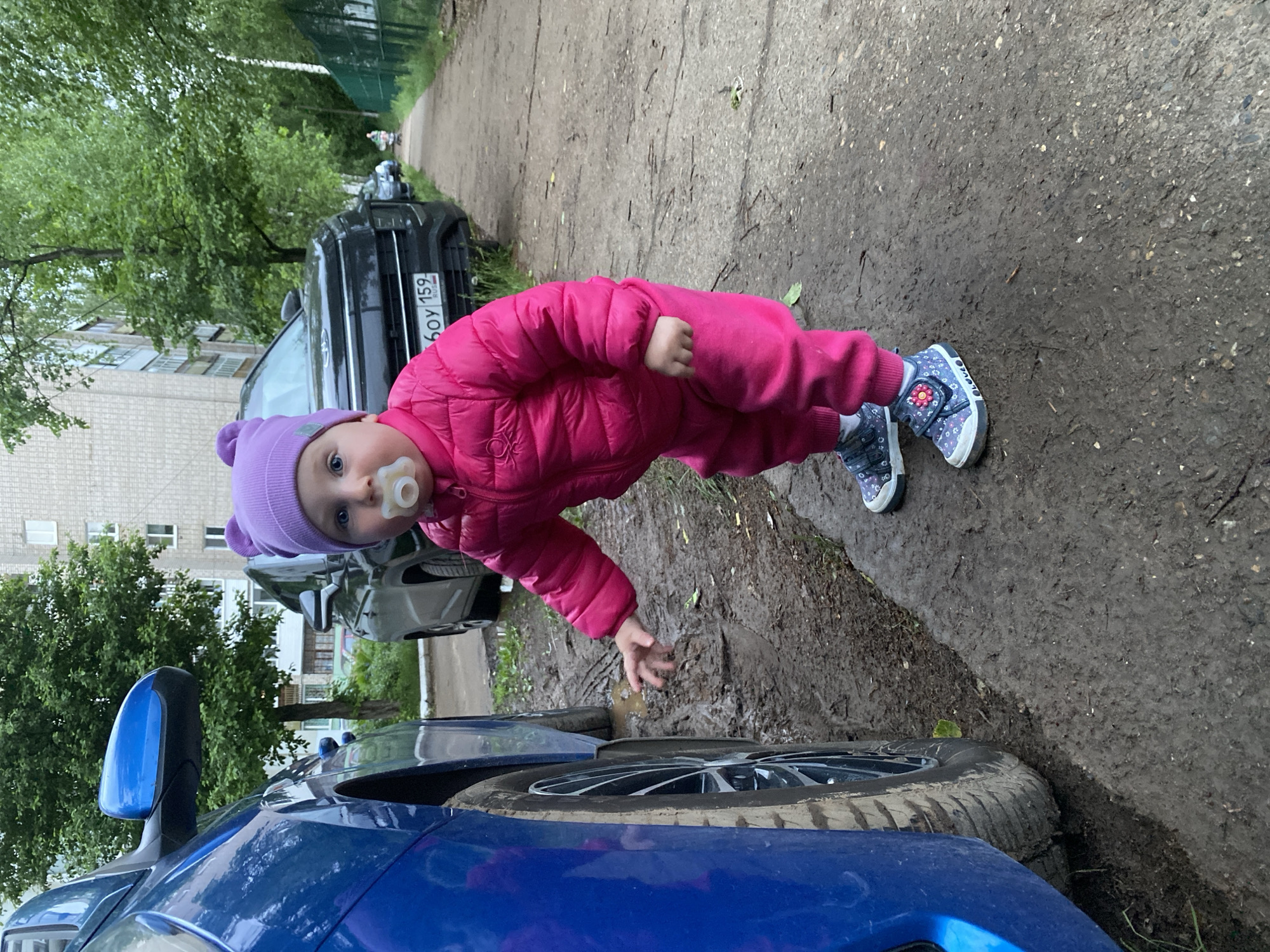 A small child wearing a pink snowsuit and a lilac hat is standing next to a blue car on a muddy, paved surface. The child is reaching towards the puddle next to the car tire. In the background are other parked cars, a building, and trees. The license plate on one of the cars reads "60У 159 RUS". The location is Chaikovsky, Russia.