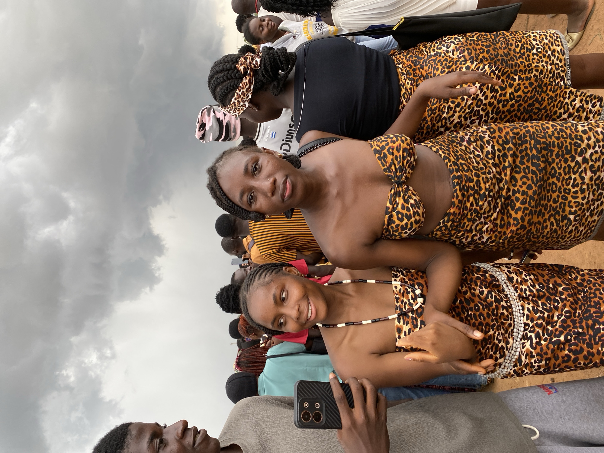 A group of people are gathered outdoors under an overcast sky. Two women in the foreground are wearing leopard-print clothing, one in a bra-top and skirt, and the other in a similarly patterned dress. The woman in the bra-top is looking at the camera. Another person holds a phone in the foreground, seemingly taking a photo of the women. Other people stand behind them. The location is identified as the city of Durimi, Nigeria.