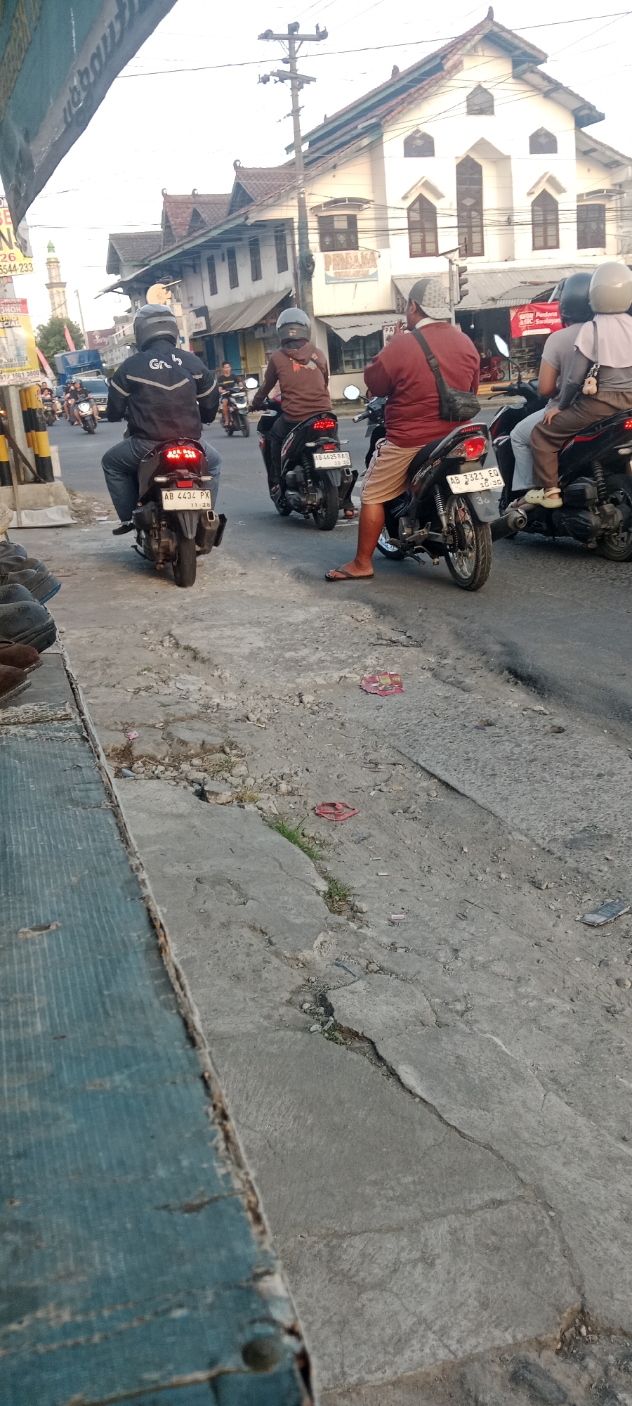 The image depicts a street scene in Maguwoharjo, Indonesia, likely during the daytime based on the lighting. In the foreground, a weathered, blue wooden structure is visible along the left edge. The street itself is paved with asphalt and concrete, showing signs of wear and tear with cracks and debris. Several motorcycles are present, with riders on them. One motorcycle in the foreground has a license plate reading "AB 4434 PX". Another motorcycle to its right has a license plate "AB 4625 BAT". Further back, more motorcycles and riders are visible, suggesting traffic. The background features buildings with traditional Indonesian architectural elements, including tiled roofs and ornate facades. A prominent white building with multiple windows and a peaked roof stands out. A minaret of a mosque can also be seen in the distance, along with a utility pole with electrical wires. The overall atmosphere appears to be that of a busy urban street.