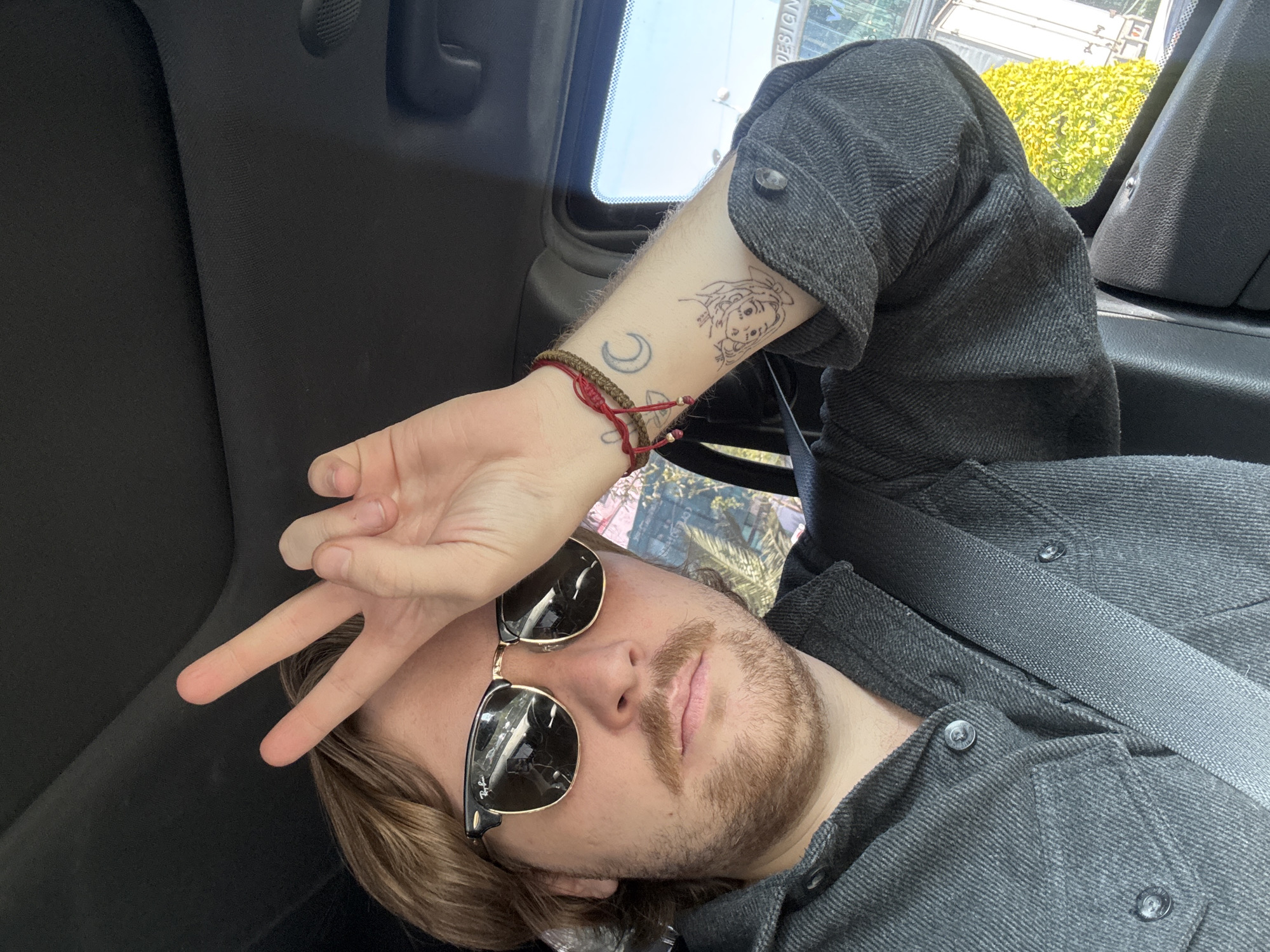 A self-portrait captured inside a vehicle, likely in Mexico City, Mexico, features a person with light brown hair, a mustache, and a beard, lying down and looking upwards. The individual wears a dark gray textured long-sleeved shirt and dark sunglasses, with a seatbelt visible across their chest.
One arm is raised, resting near the forehead, with two fingers forming a peace sign. On the visible forearm, there are two distinct tattoos: a crescent moon and a line-art drawing of a female character, reminiscent of Ariel from The Little Mermaid. The wrist is adorned with red and brown braided bracelets. The sunglasses, identified by the "Ray-Ban" logo on the frame, show reflections and appear to have some smudges or cracks on their lenses.
Through the car window, bright daylight illuminates lush green foliage, possibly a hedge or trees, along with reflections of urban structures. Part of a reversed sign with the word "DESIGN" is faintly visible. The car's interior features dark fabric on the ceiling and dark plastic panels along the side. The image conveys a casual and relaxed mood, captured during the daytime.
