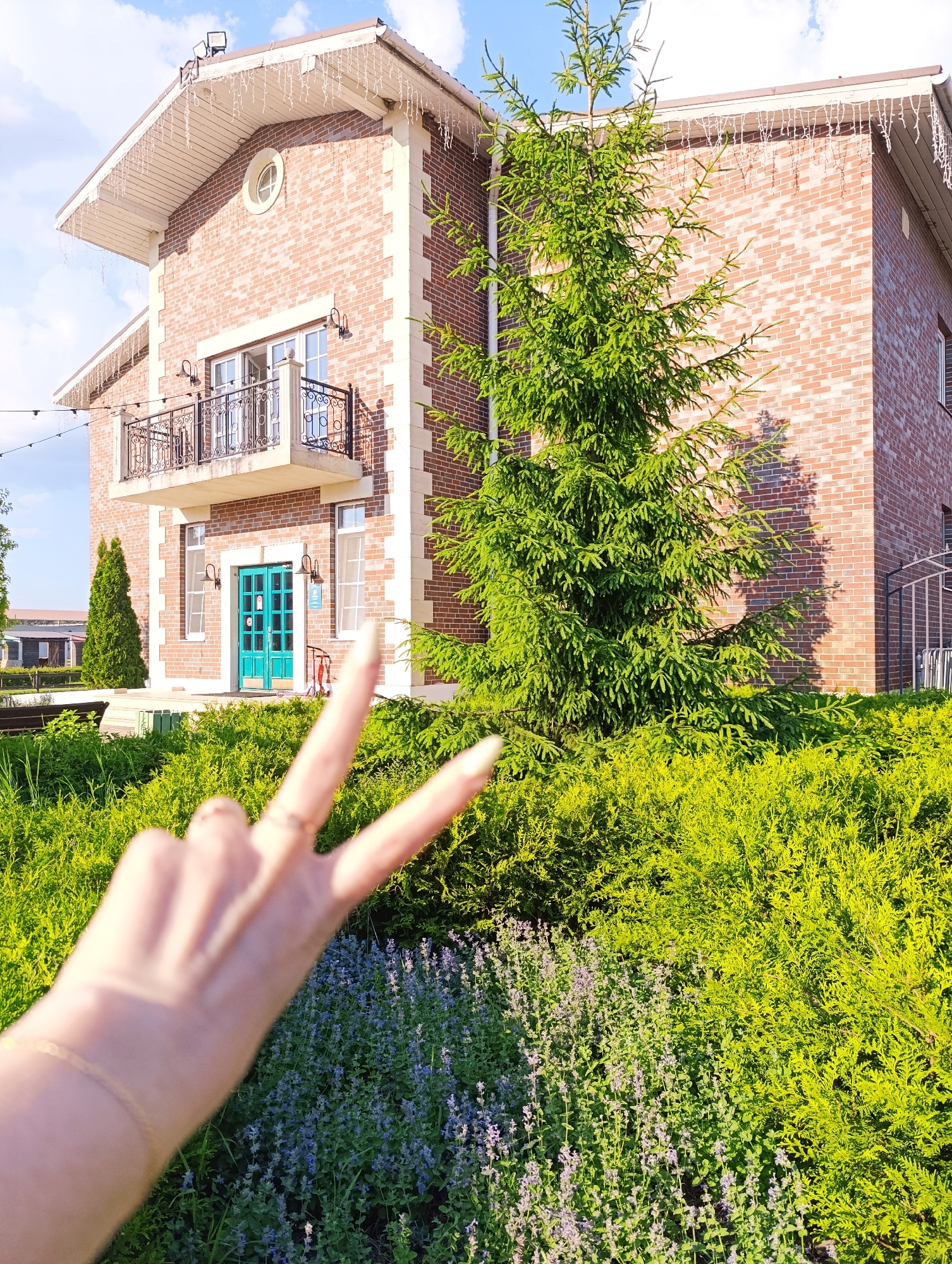 A medium shot shows a two-story brick house with a white trim roof and icicle lights hanging from the eaves. A large evergreen tree stands in front of the house, with a bed of bright green bushes and purple flowers in the foreground. In the lower corner of the frame, a hand is held up with two fingers extended in a peace sign, with a ring on one finger and a bracelet on the wrist. The scene appears to be outdoors, with a blue sky and white clouds visible in the background. The location is identified as Ivanovskoe, Russia.
