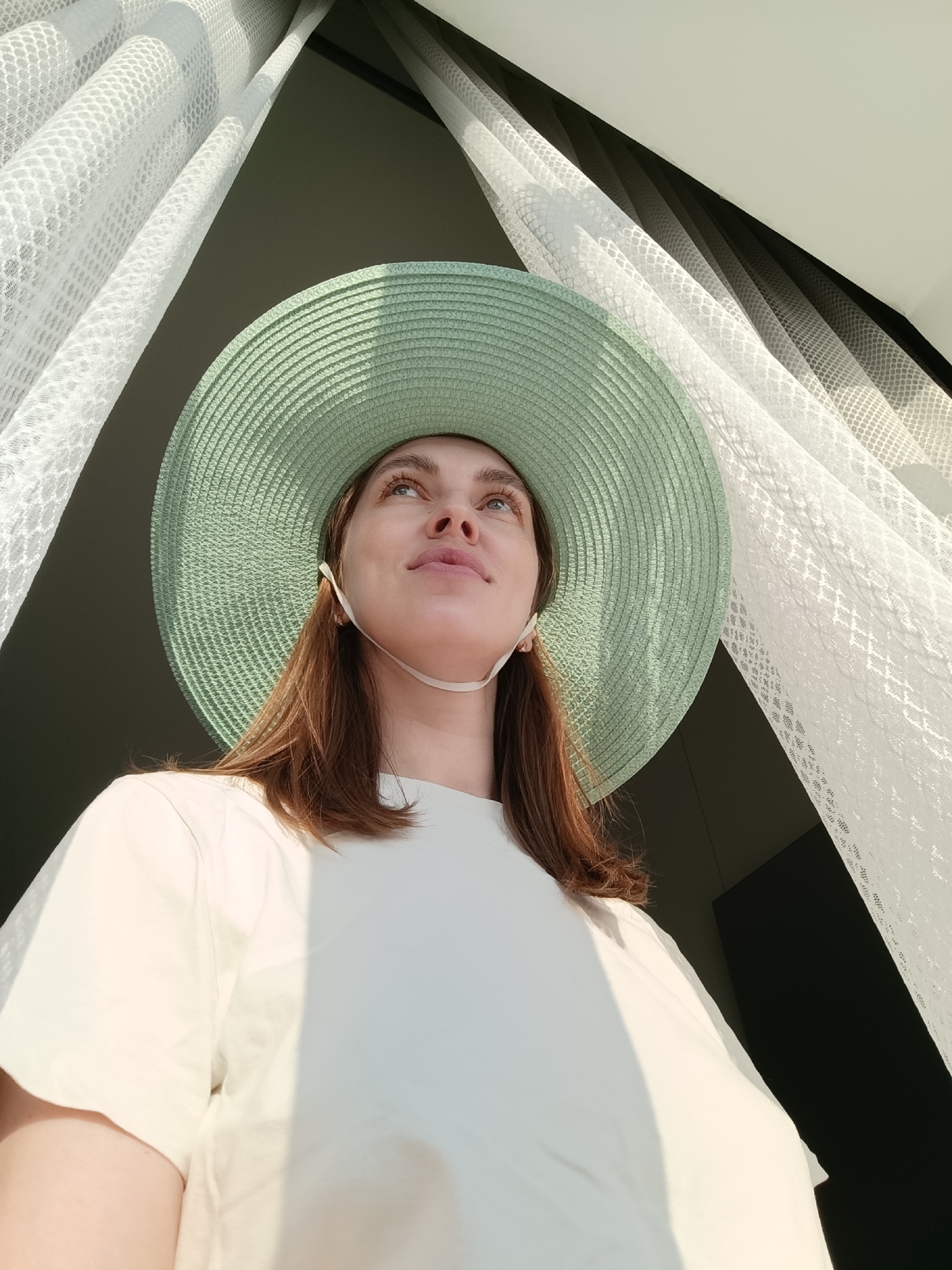A low-angle shot depicts a woman with light skin, wearing a large, light green sun hat and a white t-shirt. She is standing near a window draped with sheer white curtains. Her brown hair is shoulder-length. Her eyes are looking upwards. The background includes parts of a room, with dark elements visible beyond the curtains. The specific location within the city of Chaikovsky, Russia, cannot be determined from the image.
