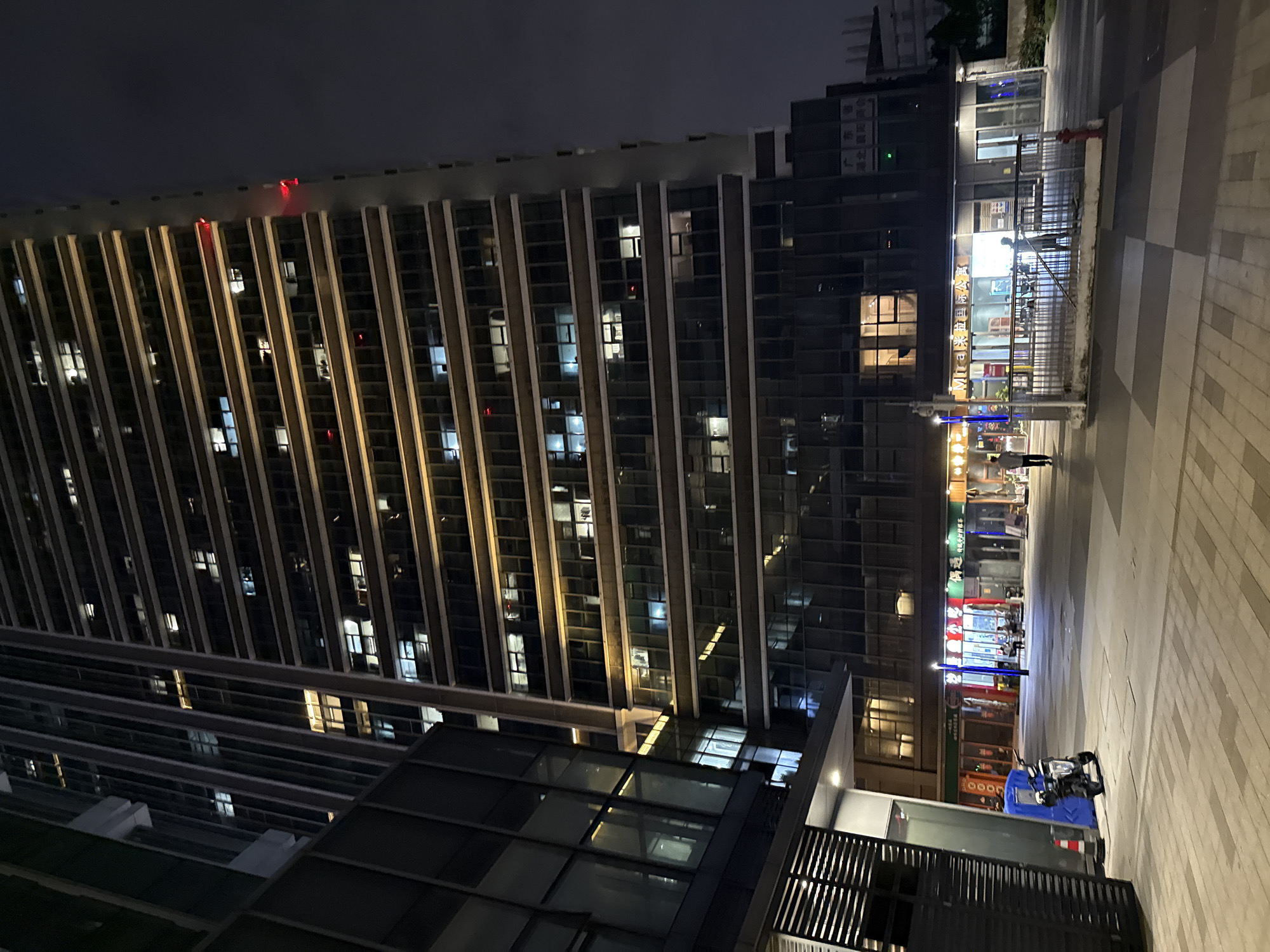 The image is a night-time, low-angle shot of a modern, multi-story building in Huangpu District, China. The building features a dark facade with numerous illuminated windows, suggesting occupancy. A portion of an adjacent building, also with dark glass windows, is visible to the left. At the base of the main building, a pedestrian area with checkered paving is present. A blue scooter is parked near a blue enclosure and an orange traffic cone. Several shops with illuminated signs are visible along the ground floor, with people present inside and outside. One sign appears to read "Mira 米拉国际公寓". A red fire hydrant is visible in the foreground. The sky is dark and overcast.
