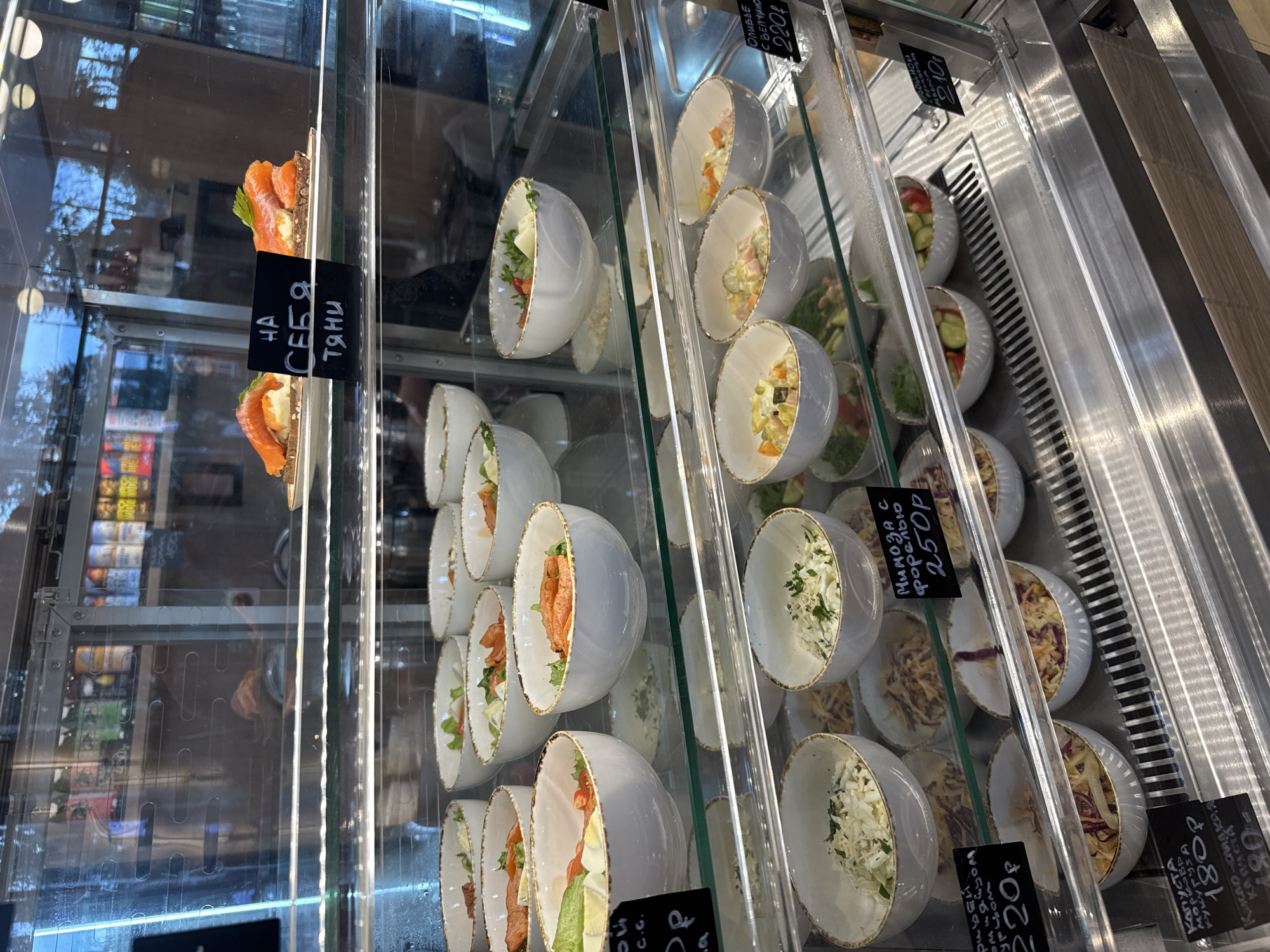 This is a medium shot, eye-level photograph of a refrigerated display case filled with various pre-packaged salads and appetizers. The display case is made of glass and stainless steel, with multiple shelves. The lighting is bright and evenly distributed, illuminating the food items.
On the top shelf, there are two small plates with what appear to be salmon appetizers, each topped with a sprig of greenery. A small black sign with white Cyrillic text is placed in front of these appetizers.
The middle shelves are filled with white ceramic bowls containing different types of salads. Some bowls contain leafy green salads with chunks of salmon, egg, and tomato. Others appear to be vegetable salads or coleslaw. Several bowls have small black signs with Cyrillic text and prices. One sign reads "МИМОЗА С ФОРЕЛЬЮ 250Р" which translates to "Mimosa with trout 250 rubles". Another sign reads "РЫБНЫЙ САЛАТ СО СПЕЦИЯМИ 220Р" translating to "Fish salad with spices 220 rubles".
The bottom shelf also contains bowls of salads, including what looks like coleslaw and other mixed vegetable salads. A sign on this shelf reads "КАПУСТА МОРКОВЬ КУКУРУЗА 180Р" which translates to "Cabbage carrot corn 180 rubles". Another sign reads "ОЛИВЬЕ С ВЯЛЕНЫМИ ТОМАТАМИ 220Р" which translates to "Olivier with sun-dried tomatoes 220 rubles".
In the background, through the glass, shelves with bottles of drinks and other items are visible, suggesting this is a store or cafe. The overall environment appears clean and organized. The specific location in the city of Yakkima, Russia cannot be determined from the image alone.