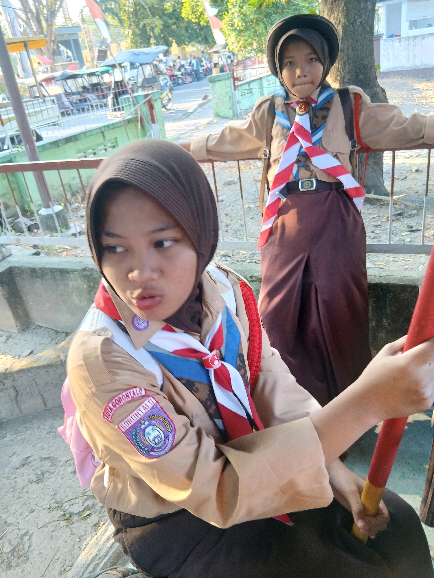 In the city of Gorontalo, Indonesia, two young girls in scout uniforms stand near a fence. The girl in the foreground wears a brown hijab and a light brown scout shirt with patches on the sleeve. She is looking to the left with her mouth slightly open. The second girl, in the background, wears a dark hat and a similar scout uniform, standing with her arms outstretched. Behind them, a street scene is visible with parked vehicles, including motorcycles and what appear to be becak (motorcycle rickshaws). Trees and buildings are also present in the background, suggesting an outdoor urban setting. The lighting indicates it is likely daytime, possibly late afternoon due to the warm tones.