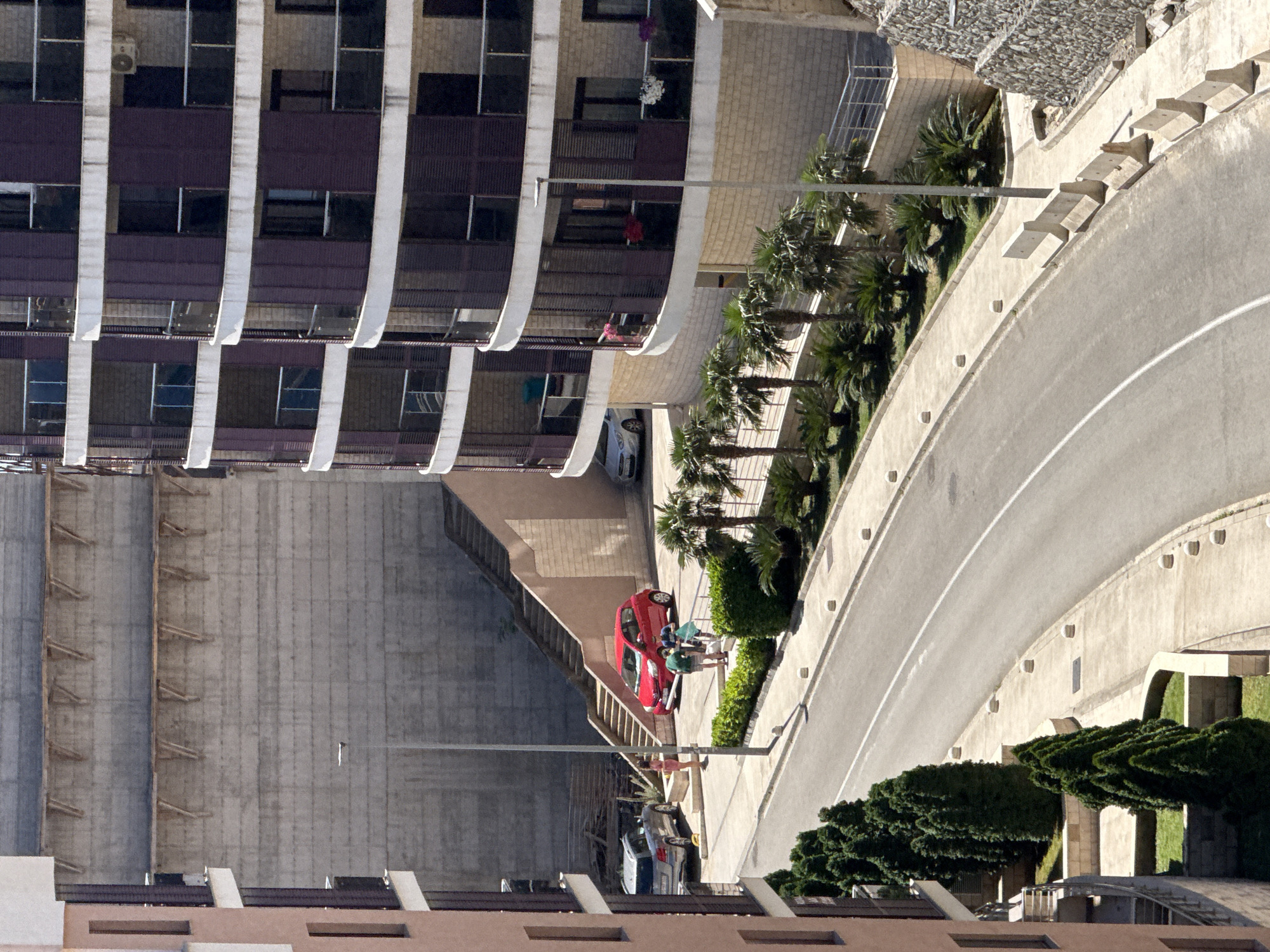 In Boreti, Montenegro, a street scene unfolds with modern apartment buildings framing a curving asphalt road. To the left, a light-colored apartment building with balconies is partially visible. Across the road, a tall concrete wall forms the backdrop for a set of concrete stairs leading upwards. A red compact car is parked at the base of the stairs, with a silver sedan beside it. Two individuals, a woman in a pink dress and a man in green shorts and a t-shirt, are standing near the red car. Another person in a blue shirt and shorts is nearby. Lush green trees and palm trees line the sides of the road, with a concrete walkway interspersed with small white bollards. Further to the right, a multi-story apartment building with curved balconies and dark shutters dominates the scene. A street lamp stands prominently in the foreground, with a concrete barrier visible along the edge of the road on the far right. The environment appears sunny and developed, suggesting a residential or commercial area.