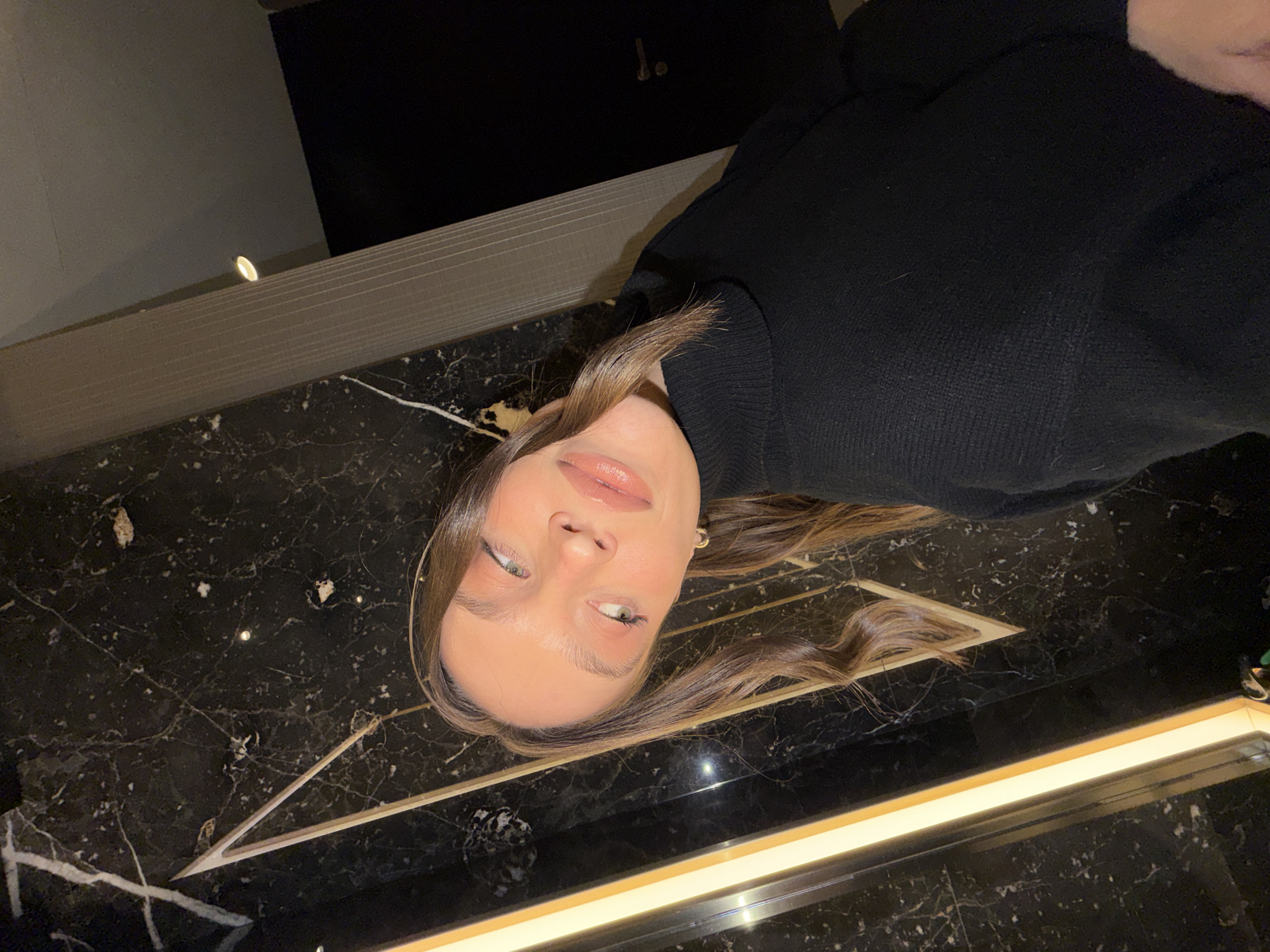 The image depicts a young adult female captured in a self-portrait, tilting her head to the left, within a modern interior space located in Budva, Montenegro. The subject has fair skin, light-colored eyes, and long, wavy brown hair that falls over her shoulders. She is wearing a black ribbed turtleneck sweater and a small gold hoop earring on her right ear. Her lips are glossed with a light pink shade. The background features dark, polished marble walls with prominent white and grey veining. A bright, warm-toned vertical light strip is integrated into the marble on the left side of the frame. A V-shaped decorative inlay, possibly metallic and gold-toned, is visible within the marble above the subject's head. To the right, a fluted light-grey column partially obscures a dark door with a visible handle. A single recessed circular light fixture is visible on the ceiling above and to the right of the subject.