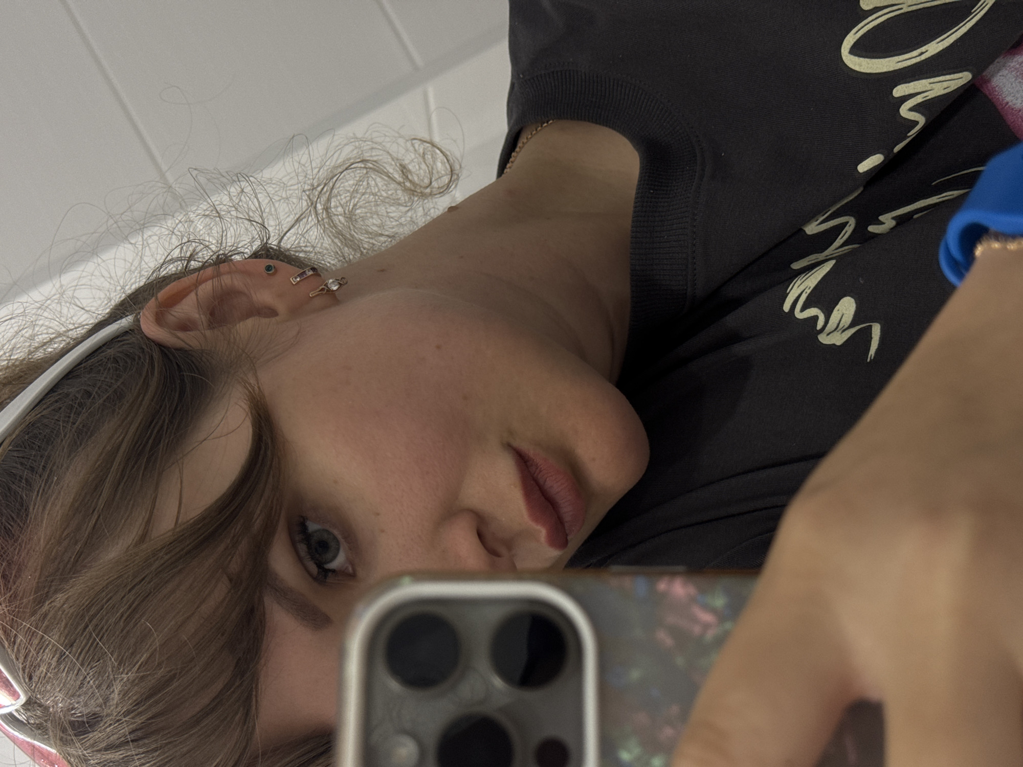 A person is taking a selfie. The person has fair skin, brown hair, and blue eyes. The person is wearing a black sweatshirt with off-white lettering. The person has multiple piercings in their left ear. The person is holding a smartphone in their right hand. The background appears to be a white surface, possibly a wall. There is no visible indication of Yevpatoriya, Ukraine.