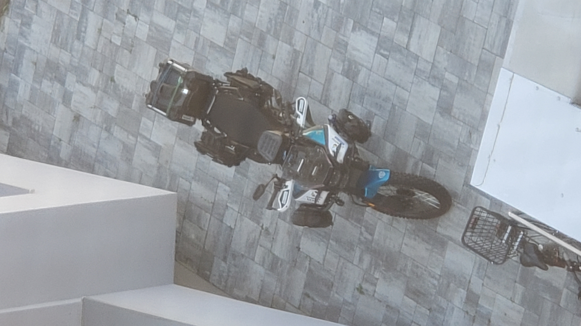 An overhead, high-angle shot captures a parked blue and white touring motorcycle on a grey tiled patio. The motorcycle is equipped with multiple black storage cases, including saddlebags and a top case, suggesting it is prepared for travel. The front of the motorcycle features a large windscreen and protective handguards. In the lower left of the frame, a black bicycle with a front basket is partially visible. The background consists of a light-colored building. The scene is static, with no visible activity. This appears to be a residential or commercial area with paved outdoor space.