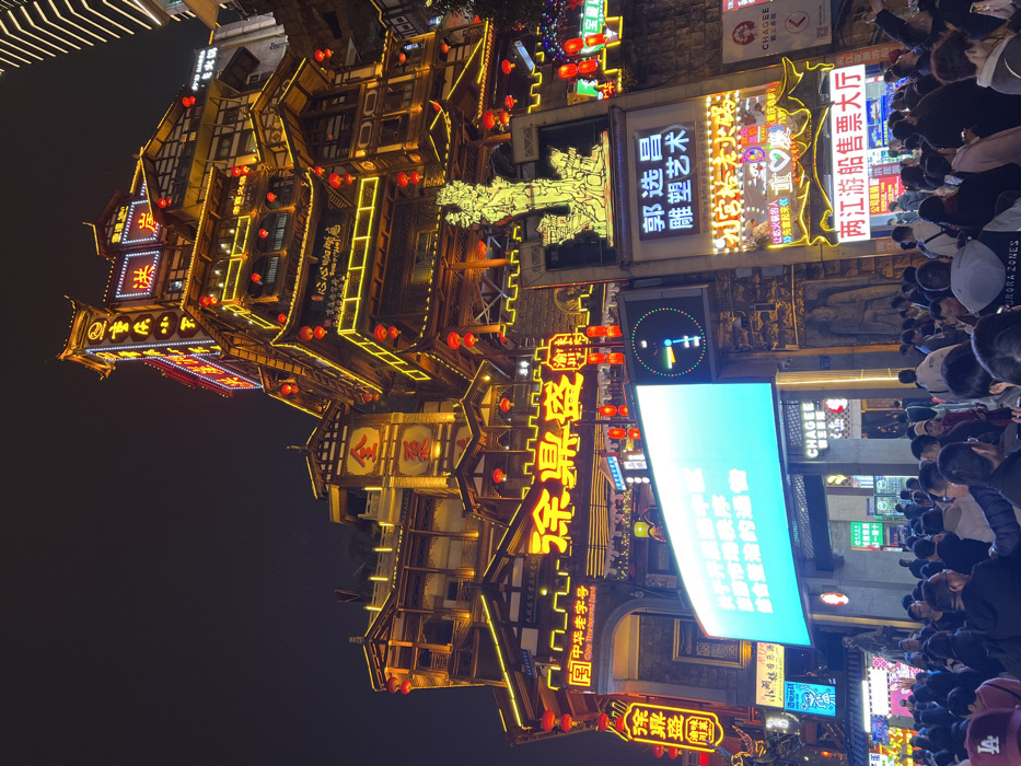 The image depicts a densely crowded nighttime street scene at Hongyadong Commercial Street in Yuzhong District, Chongqing, China. The prominent subject is a large, multi-story structure constructed in a traditional Chinese architectural style, resembling stilt houses, which dominates the upper two-thirds of the frame. This structure is extensively illuminated by warm yellow/gold neon lights outlining its various tiers and architectural features, complemented by numerous red traditional Chinese lanterns hanging from its balconies and eaves. Large Chinese characters, including "洪崖洞" (Hongyadong) and "金碧辉煌" (Golden and Brilliant), are visible in neon on different sections of the building.
At street level, a large, bright rectangular LED screen displays blue and white Chinese text, positioned above a pedestrian thoroughfare. To its right, a vertical sign features a golden, intricate sculpture-like emblem and the text "郭选昌 雕塑艺术" (Guo Xuanchang Sculpture Art). Below this, another sign reads "两江游船售票厅" (Two Rivers Cruise Ticket Office). Various other commercial signs, including "CHAGEE" and "涂鼎盛" (Tu Dingsheng), are present along the lower facades.
The lower half of the image is filled with a dense crowd of people, primarily seen from behind or as a collective mass. Individuals are engaged in walking, standing, and observing the illuminated environment. The scene is saturated with artificial light against a dark night sky, creating a vibrant, bustling urban atmosphere.