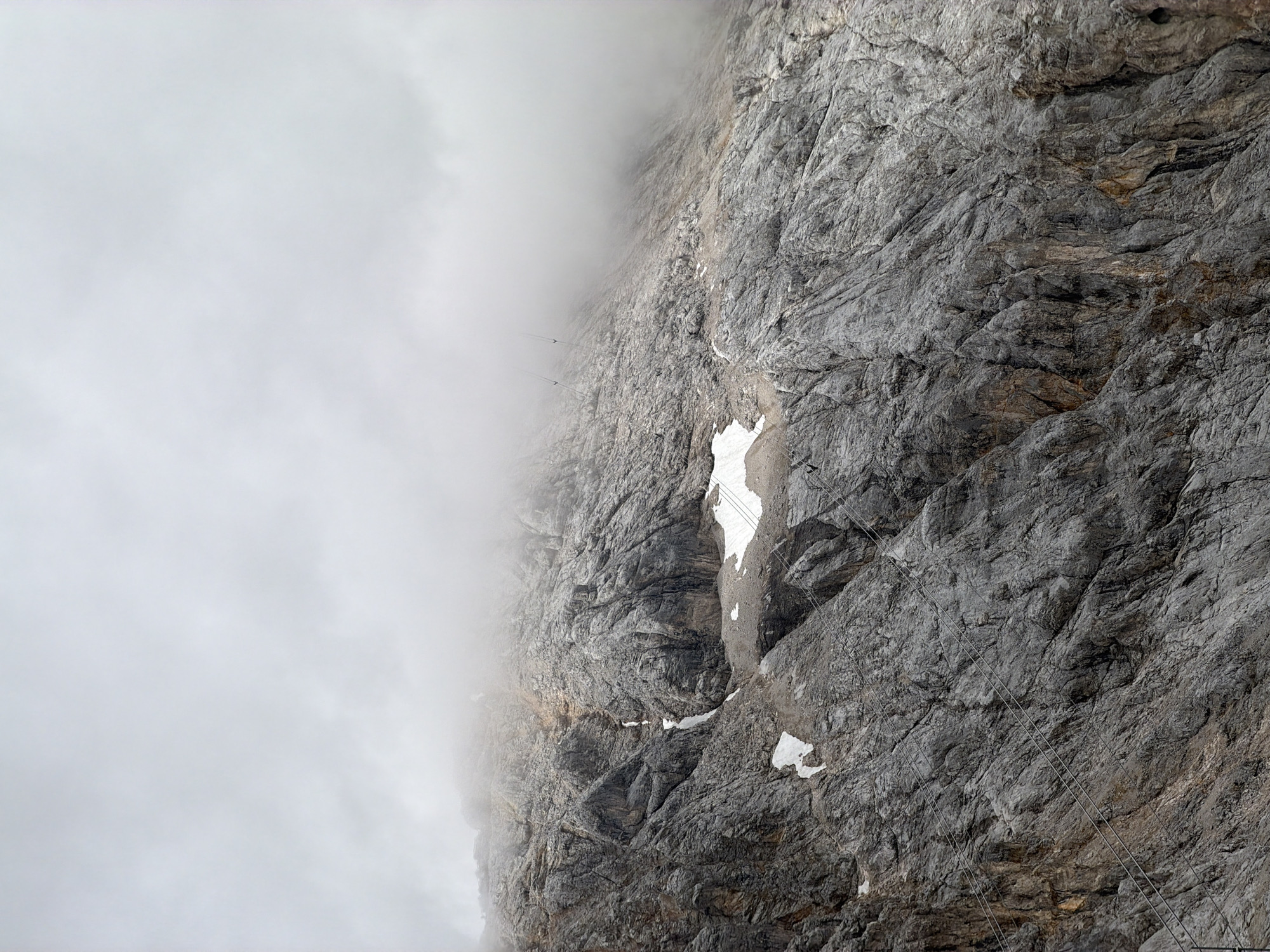 The image depicts a rugged, rocky mountainside with patches of snow. The upper portion of the image is obscured by thick, gray clouds, suggesting overcast weather or fog. Several thin, dark lines, which appear to be cables from a cable car system, are visible stretching across the rocks. In the distance, silhouetted against the sky and partially obscured by clouds, are several small, dark structures that are likely cable car cabins. The mountain's surface is deeply fissured and textured, indicating a weathered rock face. There are no visible people or animals. This scene is consistent with the Zugspitze, Germany's highest peak, located near Garmisch-Partenkirchen.