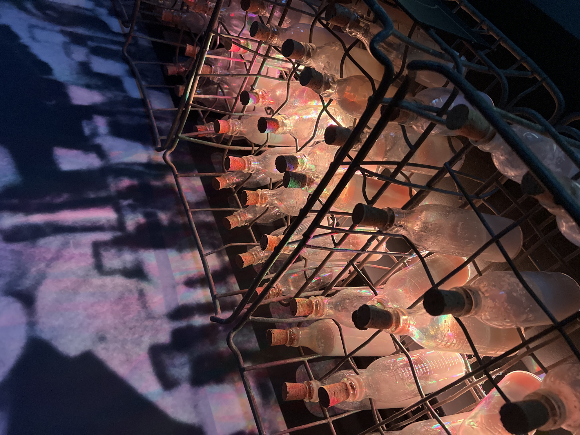 The image shows a wire rack filled with glass bottles, each sealed with a cork. Light shines through the bottles, creating a colorful, iridescent effect. The rack appears to be mounted on a wall, and a shadow or projection is visible behind it. The location is unknown.