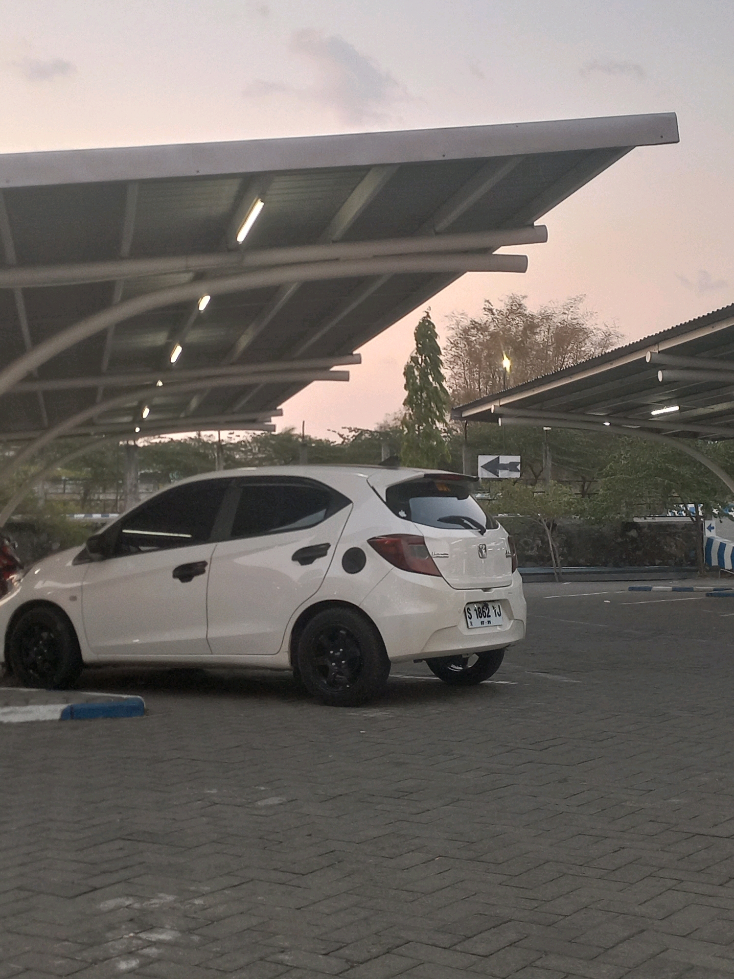 A white hatchback car is parked under a covered parking structure. The car is facing to the left, and its rear is towards the viewer. The car has black rims and tinted windows. The license plate is visible and reads "S 1862 TJ". In the background, there are trees and a sign with an arrow pointing to the left. The ground is paved with interlocking bricks. The sky is a soft pinkish hue, suggesting it is either sunrise or sunset. The lighting fixtures under the parking structure are visible and appear to be fluorescent tubes. The overall scene is one of a quiet parking area at dusk or dawn.