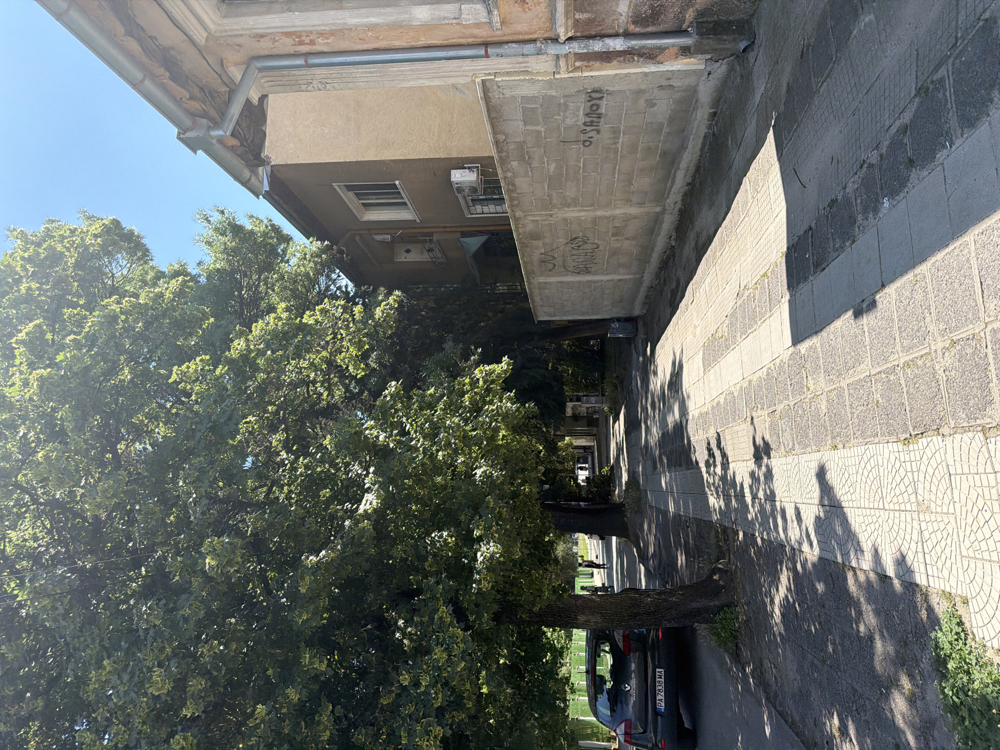 The image depicts a sun-dappled street scene in Pazardzhik, Bulgaria. A paved sidewalk runs along the right side of the street, bordered by a concrete block wall with graffiti. Adjacent to the sidewalk is a building with a tan-colored facade. On the left side of the street, a dark-colored car is parked. Large trees with dense green foliage cast shadows across the sidewalk and street. The sky is clear and blue. In the distance, two individuals are walking along the sidewalk. The scene appears to be a residential or commercial street in a moderately urbanized area.