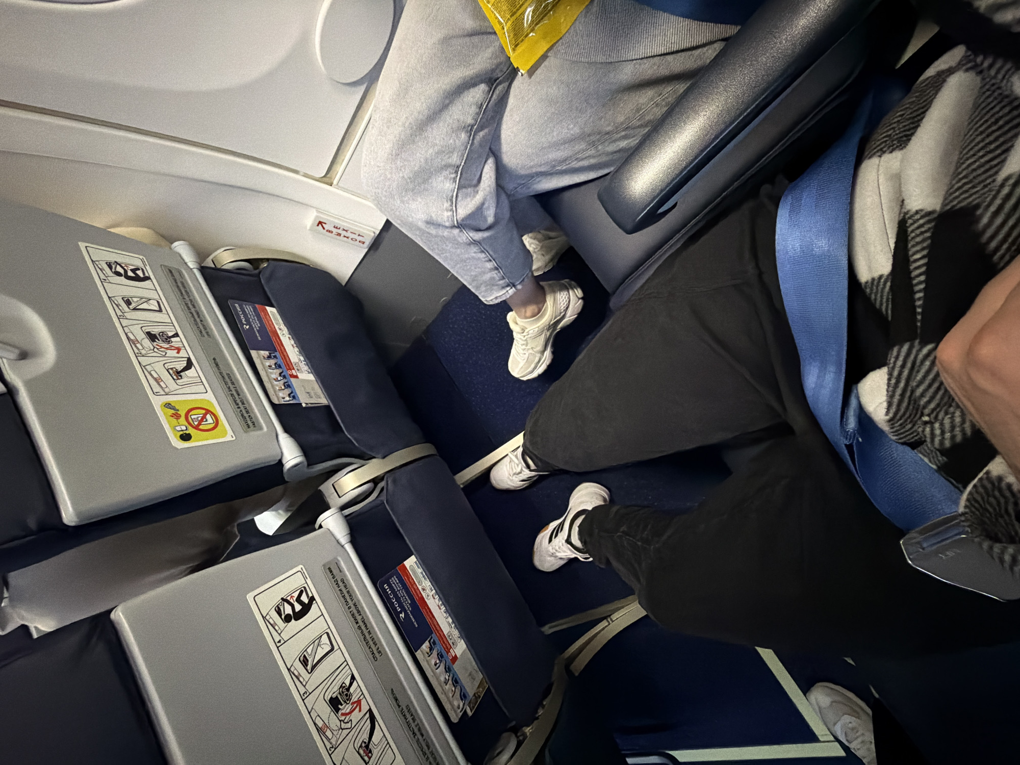 The scene depicts the interior of an aircraft cabin, taken from a first-person perspective of a seated passenger, implied to be operating in or near the city of Сокол, Russia, given the Rossiya Airlines branding. The primary subject is the legroom and seat area in front of and to the right of the photographer.
Two pairs of legs are visible: the photographer's legs extend towards the lower center-left, clad in black pants/jeans and white sneakers with black stripes (Adidas). The photographer is wearing a blue lap seatbelt over a white and black plaid garment. To the right, another passenger's legs are visible, wearing light blue denim jeans and white sneakers. A yellow wrapper or bag rests partially on this passenger's lap.
The aircraft cabin features dark blue carpeting on the floor and grey plastic seatbacks with blue fabric cushions. Each seatback is equipped with a dark blue fabric pocket containing safety information cards, one clearly displaying the "РОССИЯ" (Rossiya Airlines) logo. These cards feature illustrated instructions for seatbelt fastening, life vest usage, and electronic device restrictions, with text in both Russian and English. On the right, the aircraft's white interior wall is visible, featuring a red "ВЫХОД" (Exit) sign with an arrow pointing upwards, indicating an exit. The overall lighting is dim, suggesting an in-flight or night-time setting.