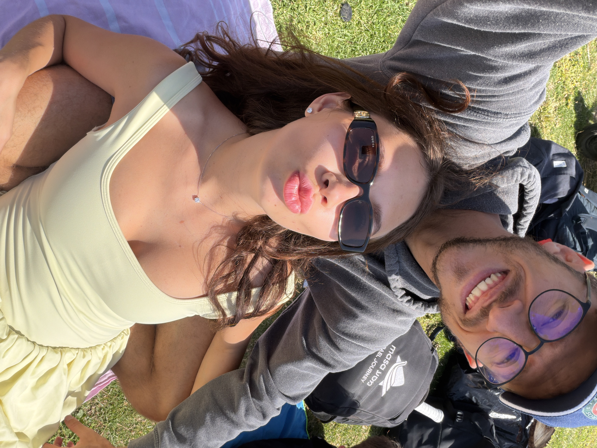 A young couple is lying on the grass in Tel Aviv, Israel. The woman is wearing a light yellow dress and sunglasses, and she has a silver necklace. The man is wearing a grey hoodie and glasses, and he has a baseball cap. They are lying on a blanket, and there are bags near them. The grass is green, and there are trees in the background.