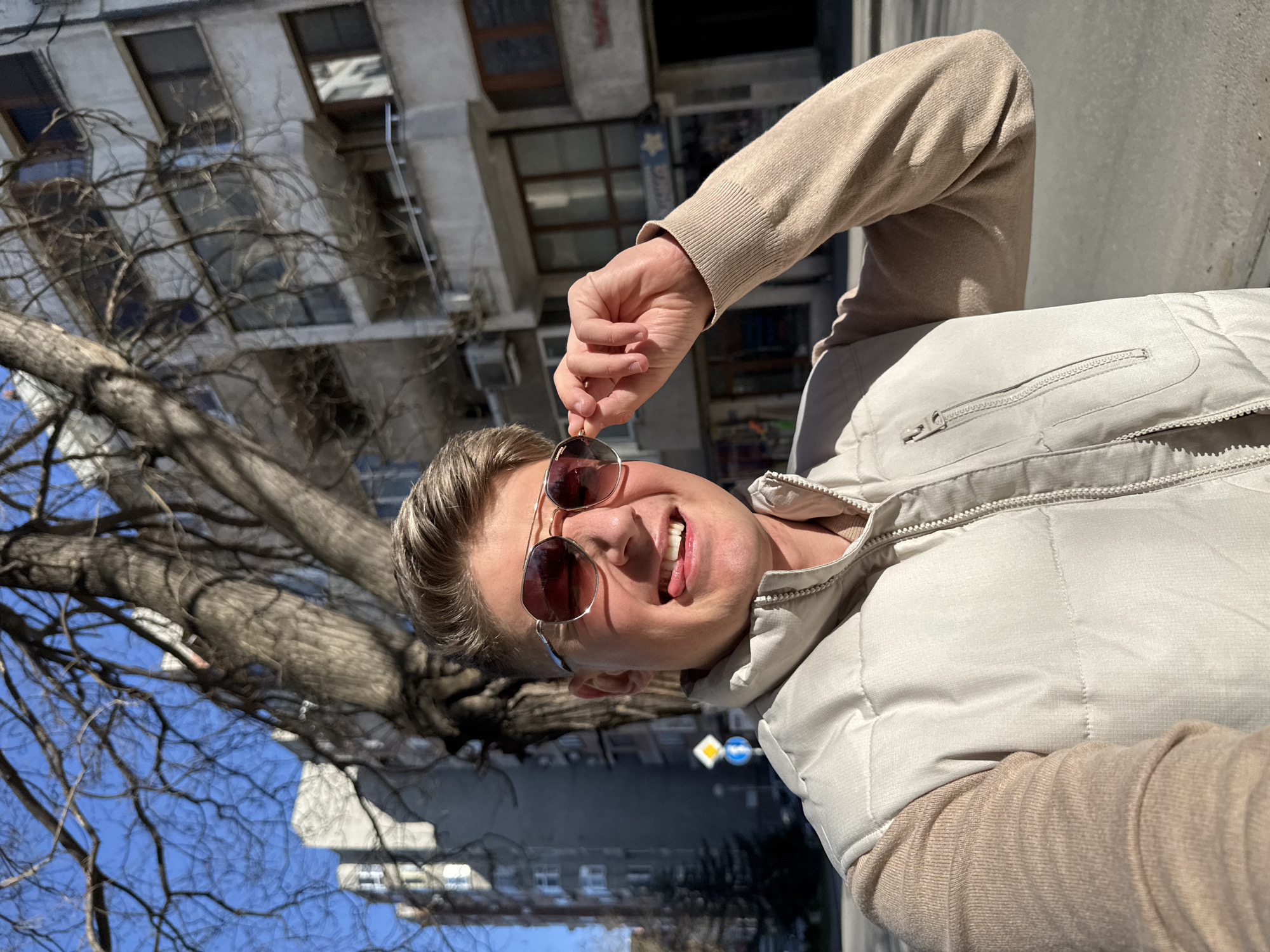A man stands outside a building in the city of Plovdiv, Bulgaria. He is smiling and holding sunglasses with a gold frame, his tongue sticking out. He is wearing a beige sweater and a sleeveless beige jacket. Behind him are buildings, a bare tree, and street signs.