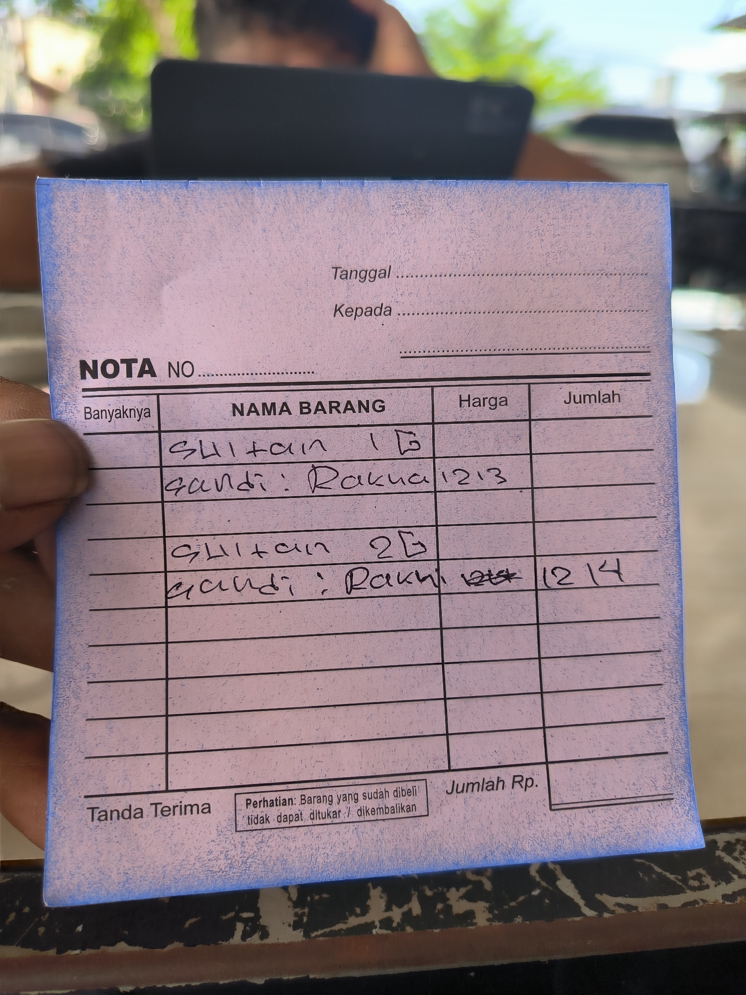 The image shows a hand holding a blue receipt on a table. The receipt is partially filled out with handwritten notes in Indonesian. The receipt lists items "Sultan" and "Gandi: Rakua 1213" and "Sultan 2G" and "Gandi: Rakhi". The number 1214 is written in the column "Jumlah" for the second set of items. The text "NOTA NO" is at the top of the receipt. The receipt also contains "Tanggal", "Kepada", "Harga", "Jumlah", "Tanda Terima", and "Jumlah Rp." The text "Perhatian: Barang yang sudah dibeli tidak dapat ditukar / dikembalikan" is at the bottom of the receipt. In the background, there is a blurry scene with greenery, a dark laptop or tablet, and a person. The setting is Lhokseumawe, Indonesia.