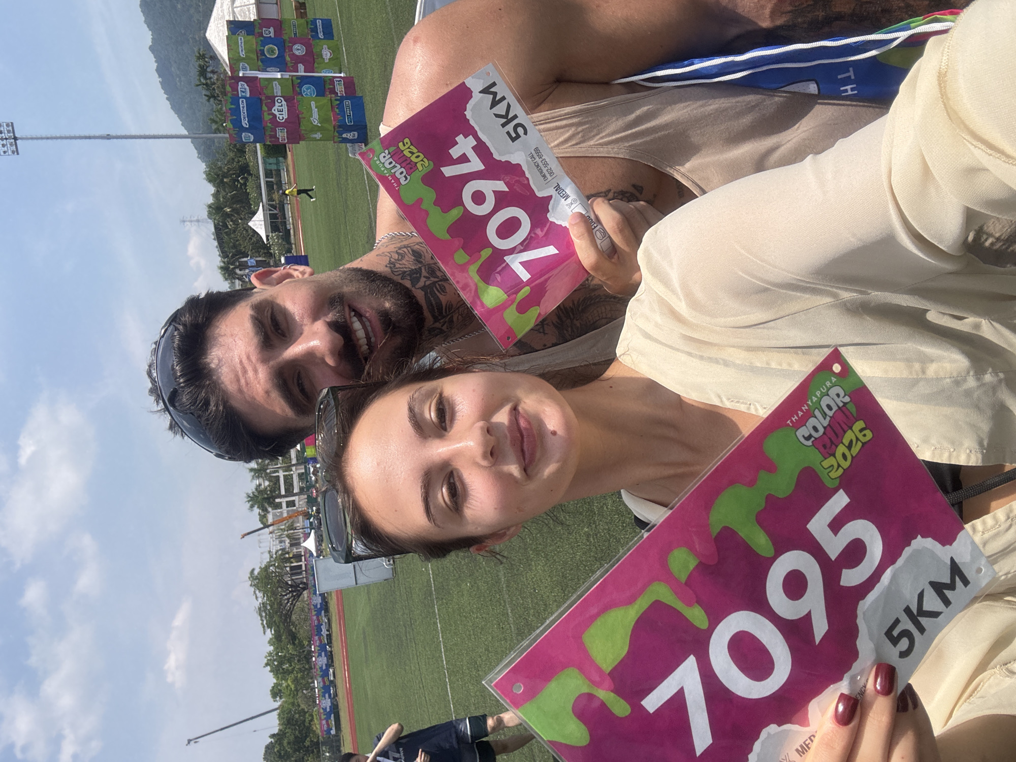 Here is a description of the image:
Two people, a man and a woman, are posing in a selfie on what appears to be a sports field. Both are holding up bibs from the "Color Run 2026", with the number "7095" on the woman's bib and "7094" on the man's. The bibs also indicate that the race is a 5KM run in "Thanyapura". The man is wearing a tank top and sunglasses and has tattoos. The woman also has sunglasses perched on her head and is wearing a light-colored top.
In the background, there is a sports field, light posts, and a sign that says "DECATHLON KASY". There are some buildings and trees in the distance and a clear sky with a few clouds. The location is likely in Thanyapura, Thep Krasatti, Thailand, as indicated by the bibs.