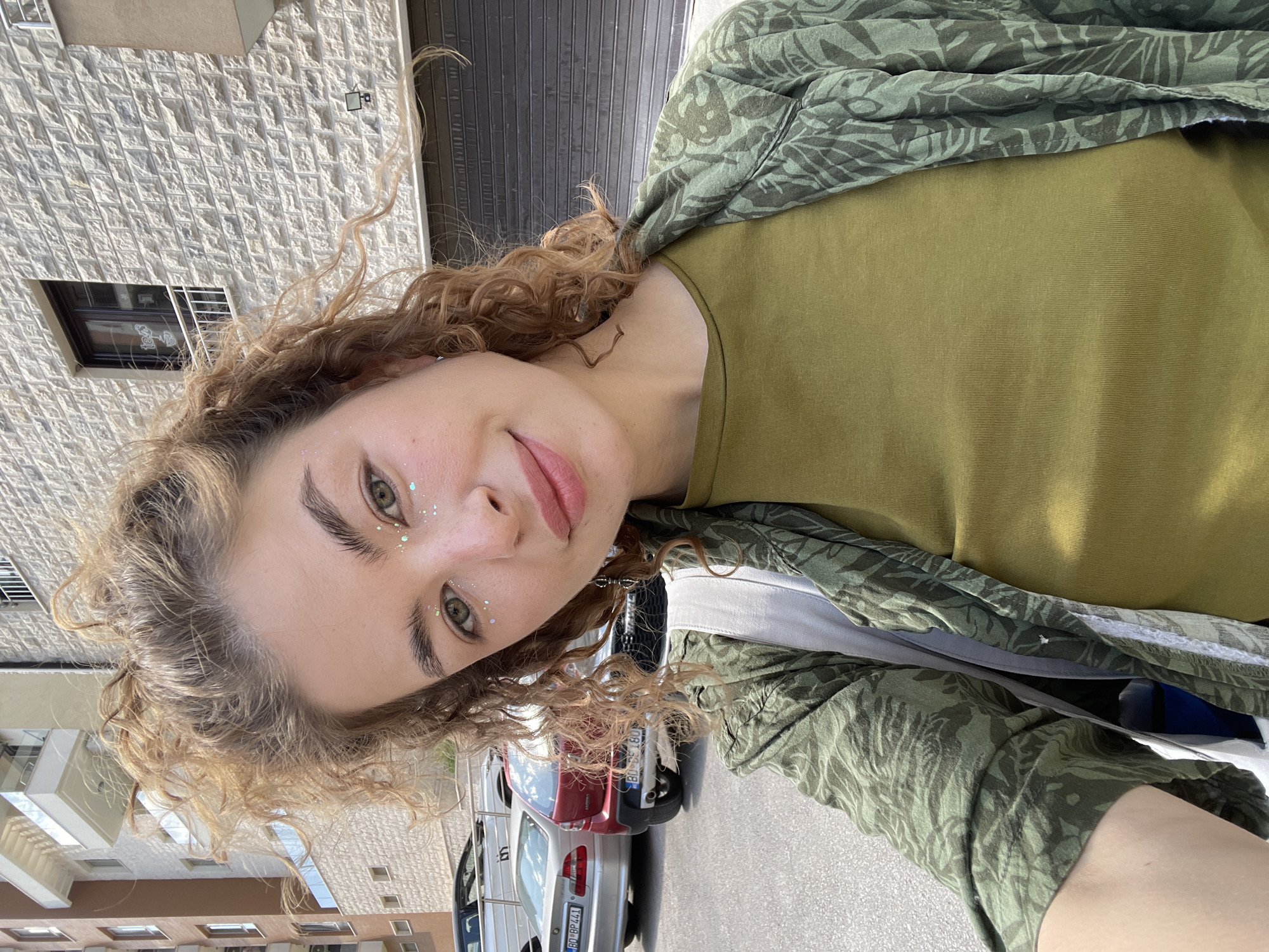 A young woman with curly blonde hair and glitter on her face is taking a selfie in Boreti, Montenegro. She is wearing a green t-shirt and a green patterned jacket. Behind her are parked cars and buildings. The location appears to be a street.