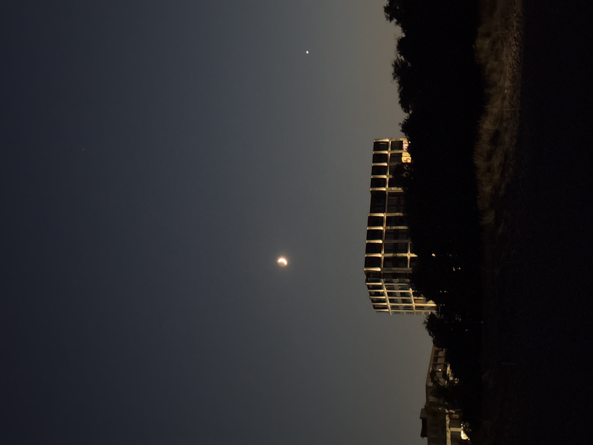 The image depicts a twilight scene over the city of Burgas, Bulgaria, with a crescent moon prominently visible in the dark blue sky. To the right of the moon, a bright star or planet is visible. In the midground, a modern building with a distinctive grid-like facade is illuminated from within. Silhouetted trees and other, less distinct buildings form a dark band across the lower portion of the image, obscuring the ground level and any potential human activity. The overall impression is one of an urban landscape transitioning from day to night, marked by celestial bodies and architectural structures.