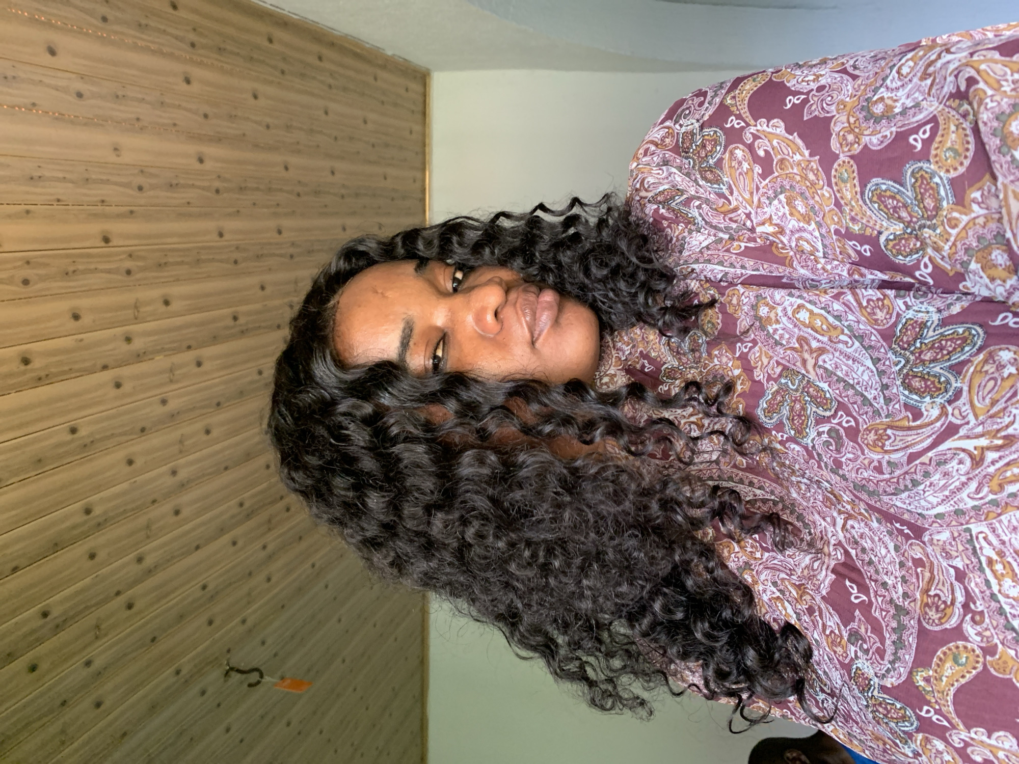 In Barakin Akawo, Nigeria, a woman with long, dark, curly hair is captured in a close-up, waist-up portrait. She is wearing a patterned garment in shades of burgundy, pink, and cream, featuring an intricate paisley design. Her gaze is directed towards the viewer, with a slight tilt of her head. The background consists of a paneled wooden ceiling, with a hook and an orange tag visible in the upper left corner. To the right, a white wall meets the wooden ceiling, and an arched doorway is partially visible. The lighting suggests an indoor setting, possibly during the day. In the far left background, a person wearing a blue shirt can be seen, their face obscured.