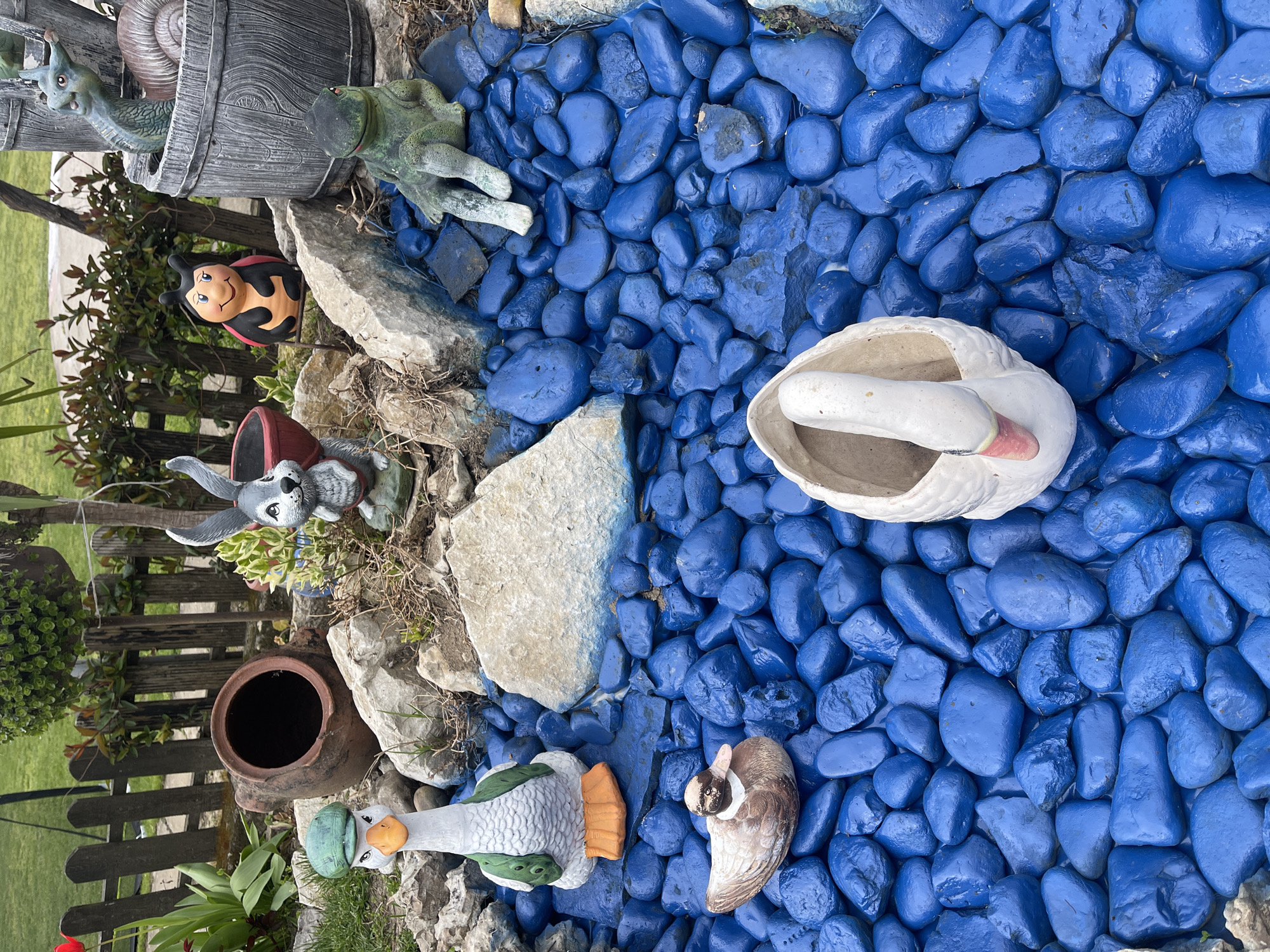 A garden decoration is seen from an overhead perspective. A collection of garden ornaments and decorations are arranged among large, intensely blue-colored stones, creating a pond-like illusion. Elements include a barrel containing a snail and a frog, a ladybug statue, a rabbit, two ceramic ducks, a white swan planter, and an earthen pot. It is potentially located in Somovit, Bulgaria.