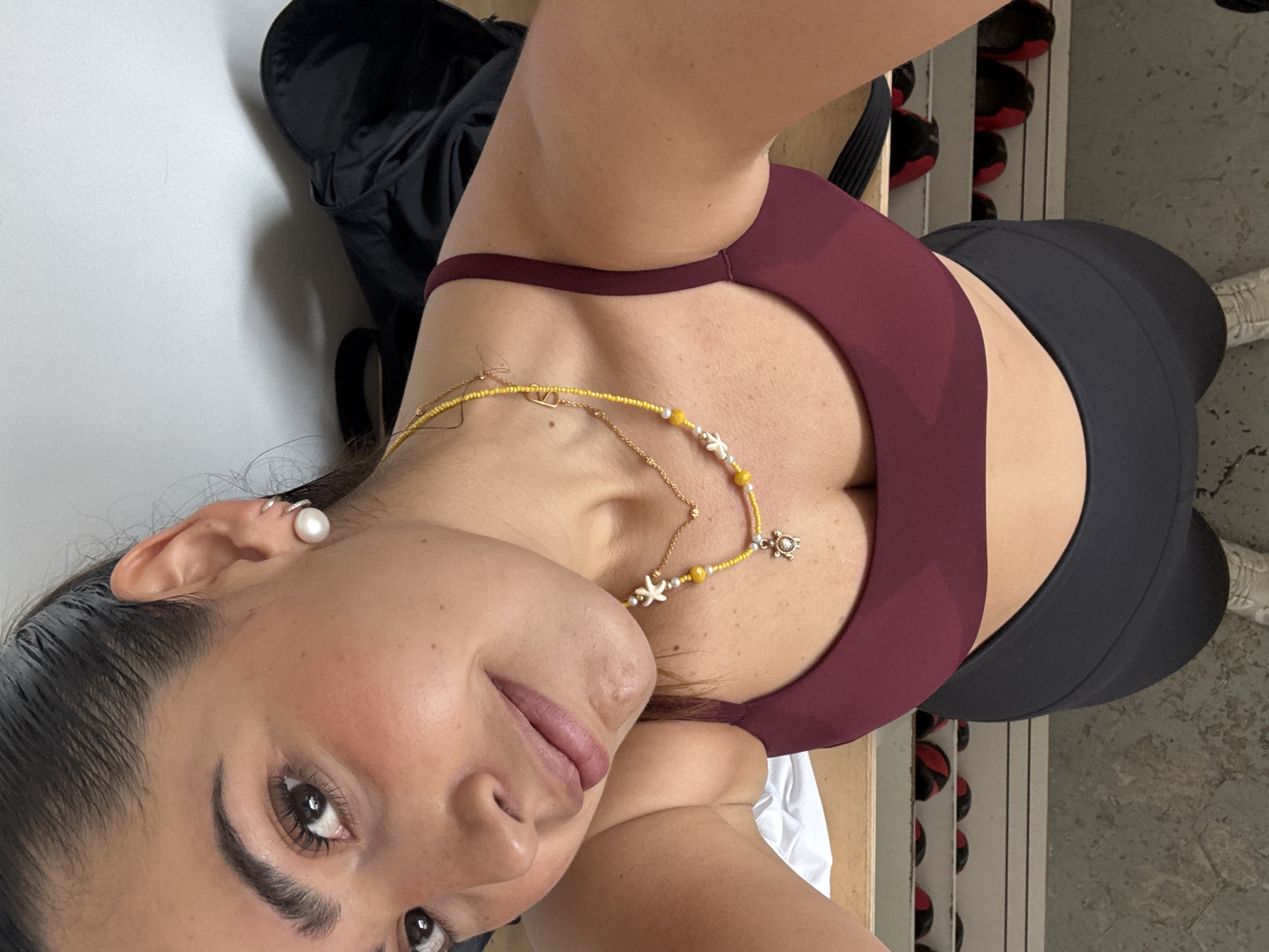 The image is a selfie taken from a high angle looking down at a woman. She has fair skin, dark hair pulled back, and brown eyes. She is wearing a maroon sports bra and black leggings. Around her neck are two gold necklaces, one beaded and one with pendants shaped like sea creatures. In her left ear is a large pearl earring.
Behind her is a black backpack on a white surface, possibly a bench or table. To the right, there is a rack with exercise balls, and concrete flooring is visible. The location, Puebla, Mexico, is not discernible from this image.
