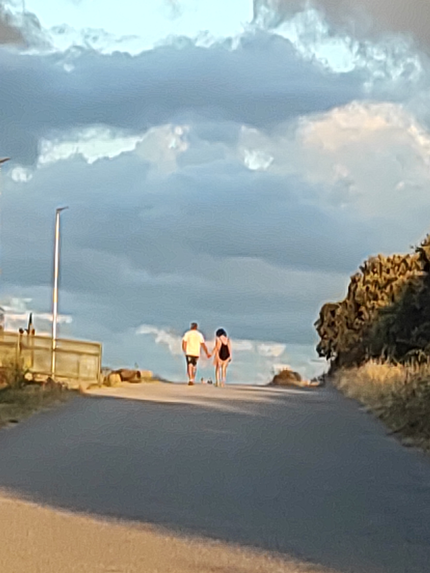 A couple walks hand-in-hand down a paved path. The male is wearing a white t-shirt and dark shorts, while the female wears a light-colored one-piece bathing suit. They are walking away from the camera towards a backdrop of a cloudy sky and a tree-lined embankment. To the left, a modern streetlamp stands near a low wall and some sandy vegetation. The scene appears to be a coastal or beachfront area.
