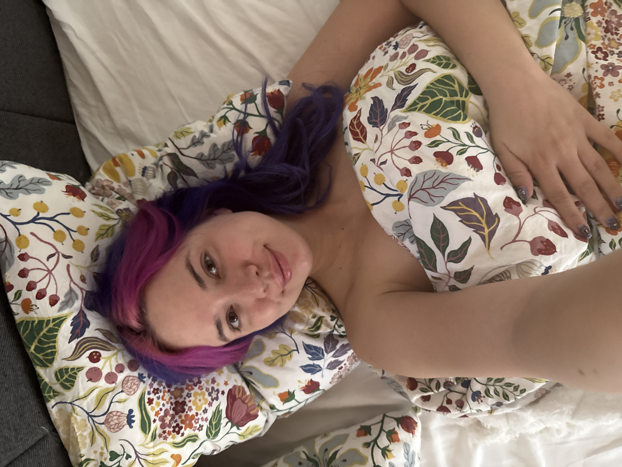 A person with purple and pink hair is lying in a bed with floral print sheets and pillowcases. The bed is on a grey couch, and the sheets are white. The location cannot be verified based on the image.