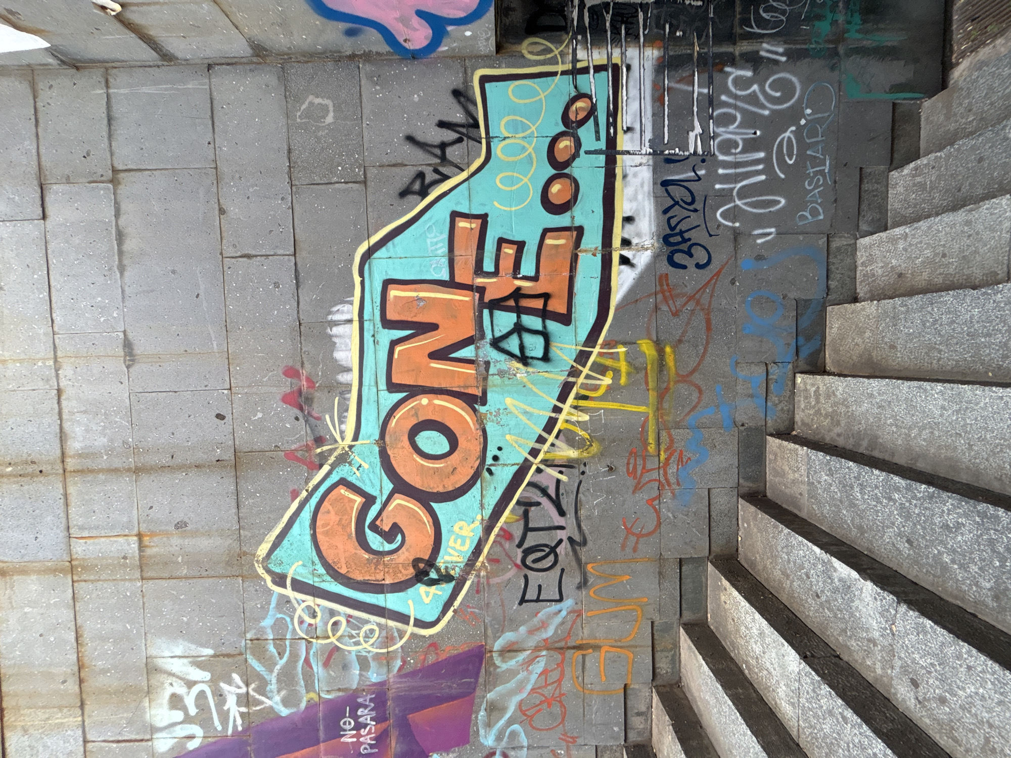 The image shows a section of an outdoor urban environment, likely a passageway or underpass, characterized by concrete block walls and a set of granite stairs. The ground is covered in grey concrete blocks, some of which are marked with graffiti. The graffiti art is the primary subject, featuring colorful and stylized lettering, abstract shapes, and tags.
Dominating the center of the image is a large graffiti piece in a distinctive shape, primarily filled with teal and orange colors. The letters appear to spell out "GONE," rendered in a bold, blocky style with yellow and black outlines. Within this piece, there are decorative elements such as yellow swirls and orange circles.
To the right of this main graffiti, a set of stone stairs ascends, appearing to be made of rough-textured granite. The wall to the right of the stairs also displays graffiti, including white lettering that seems to say "BASTARD" and other indecipherable tags. Other graffiti in various colors, including blue, red, and yellow, are scattered across the concrete walls, suggesting a history of artistic expression in this location.
The lighting suggests it is daytime, with shadows cast by the stairs. The overall impression is one of a gritty, urban space that has been transformed by street art. There is no clear indication of people or animals, and the scene appears to be captured from a high vantage point, looking down onto the ground and stairs.
Visible text includes: "GONE", "BASTARD", "NO- PASARA", and other illegible tags and lettering.