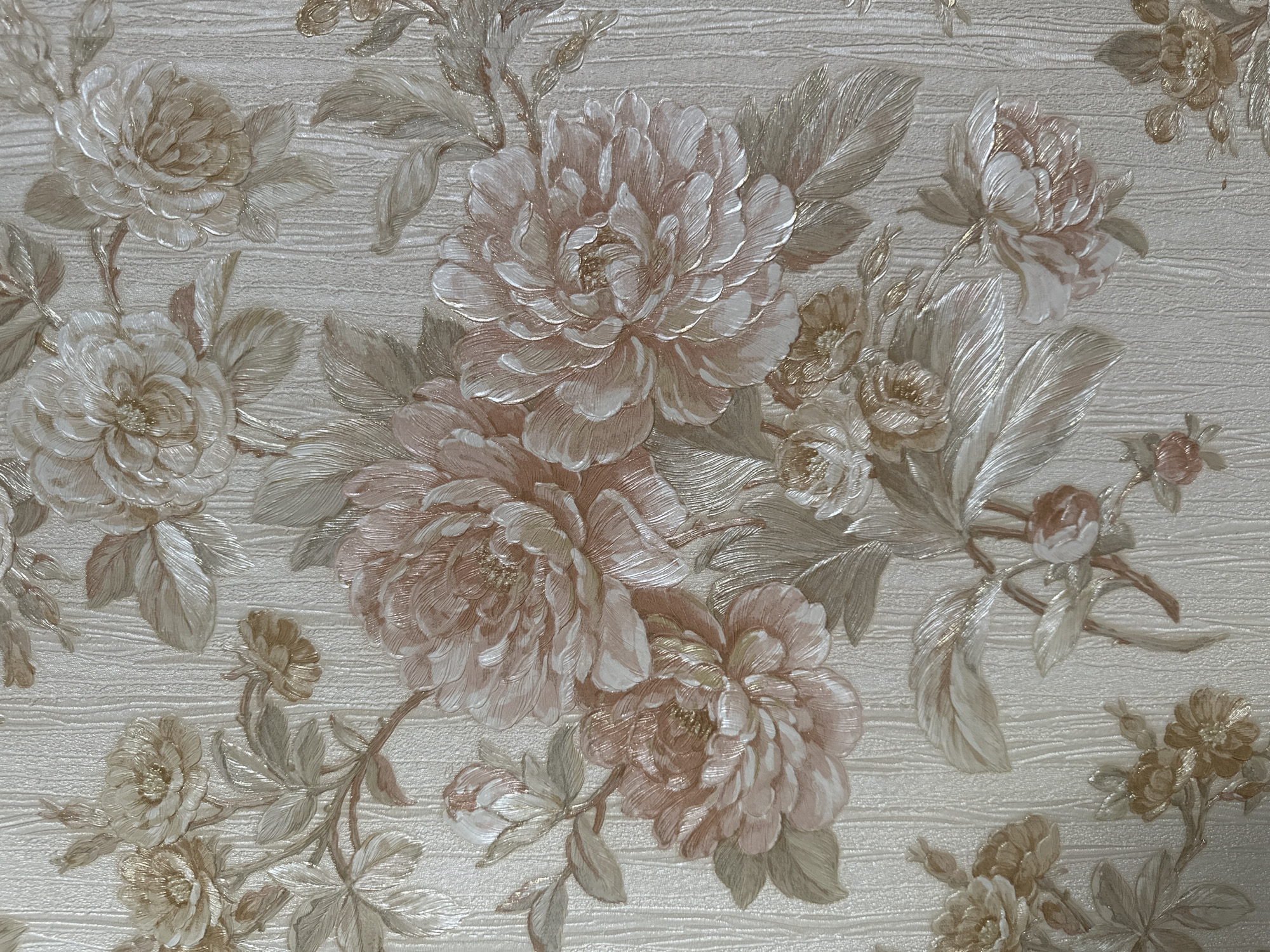 The image displays a close-up of wallpaper featuring a floral pattern. The flowers are large and appear to be peonies or similar blooms, rendered in shades of pink, cream, and gold, with textured detailing on the petals. Accompanying the flowers are various leaves and stems, also textured, in shades of green, beige, and gold. The background of the wallpaper has a subtle vertical ribbed texture. There are no people, actions, or interactions visible in this image, and it does not depict any location in Taraz city administration, Kazakhstan.