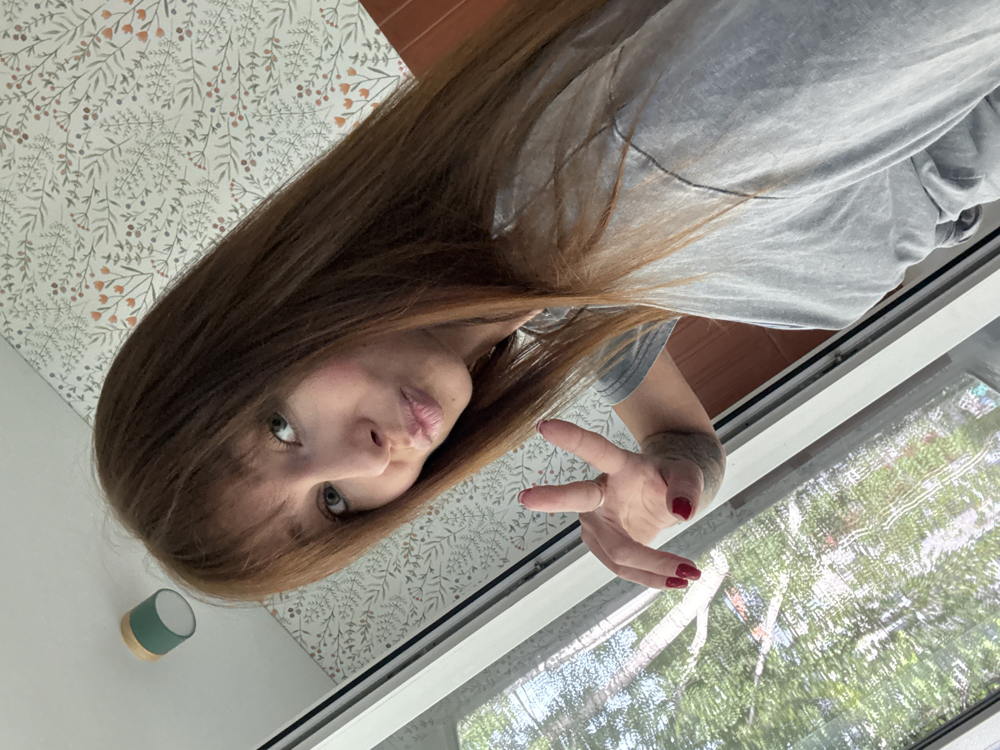 A woman with long, auburn hair is captured in a selfie, her face tilted towards the camera. She is wearing a light gray t-shirt. Her expression is playful, with her lips pursed and one eye looking directly at the camera. Her left hand is raised, with two fingers extended in a peace sign. Her nails are painted red. Behind her, a wall with a floral wallpaper pattern is visible. To the right, a window shows a reflection of trees and greenery, suggesting an outdoor setting. A small, cylindrical object with a green top and wooden base is mounted on the wall to the left of the woman. The lighting suggests it is daytime. The scene appears to be indoors, near a window. There is no visible text in the image.