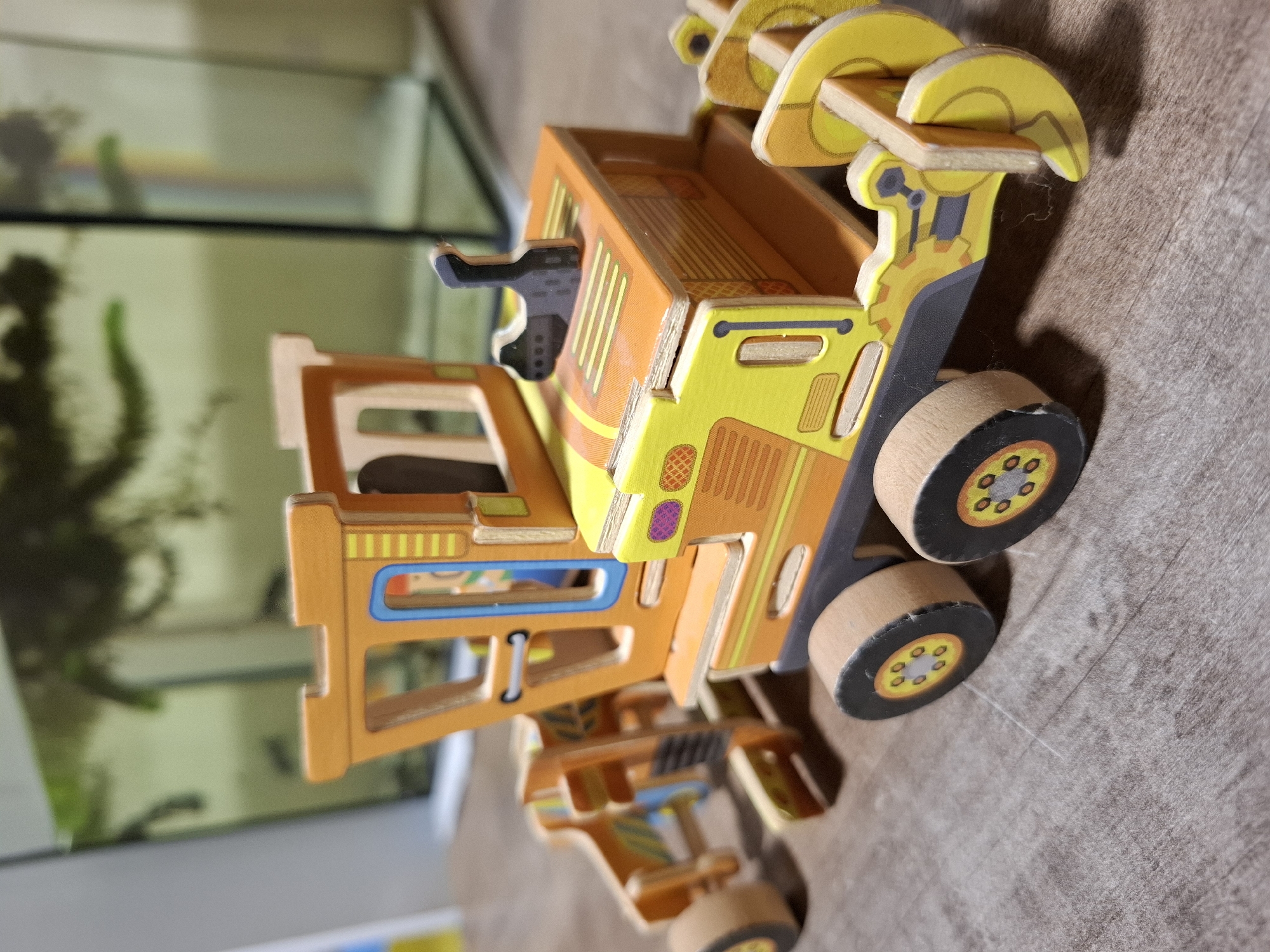 A wooden toy grader, assembled from colorful pieces, rests on a wooden surface. The toy is predominantly orange and yellow, with blue and black accents. It features wheels, a cab, and mechanical arm attachments. In the background, an aquarium with aquatic plants and what appear to be fish is visible, with a blurry green and brown environment beyond. The image is taken from a slightly elevated angle, focusing on the toy.