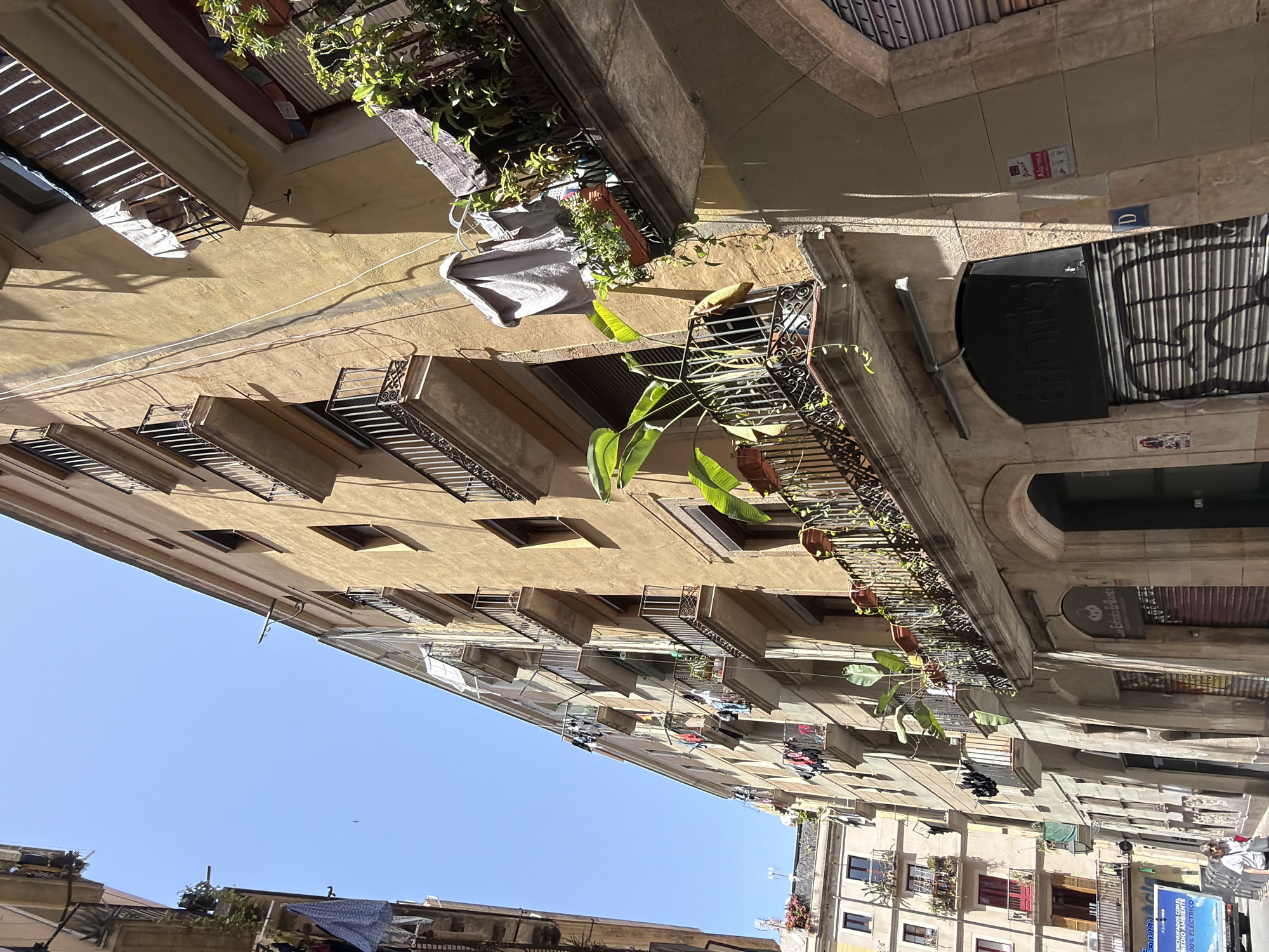 The image shows a street scene in Barcelona, Spain, with a focus on the architecture and daily life. The buildings are old, with yellow facades, balconies, and wrought-iron railings. Laundry is hanging out to dry from several balconies, a common sight in residential areas. Potted plants are also visible on the balconies, adding greenery to the urban landscape. The street is narrow, with shadows cast by the buildings, indicating a sunny day. In the foreground, a portion of a store with a closed metal shutter is visible. A woman is walking on the sidewalk. A white truck with Spanish text is parked on the street. The overall impression is of a vibrant, lived-in urban environment.