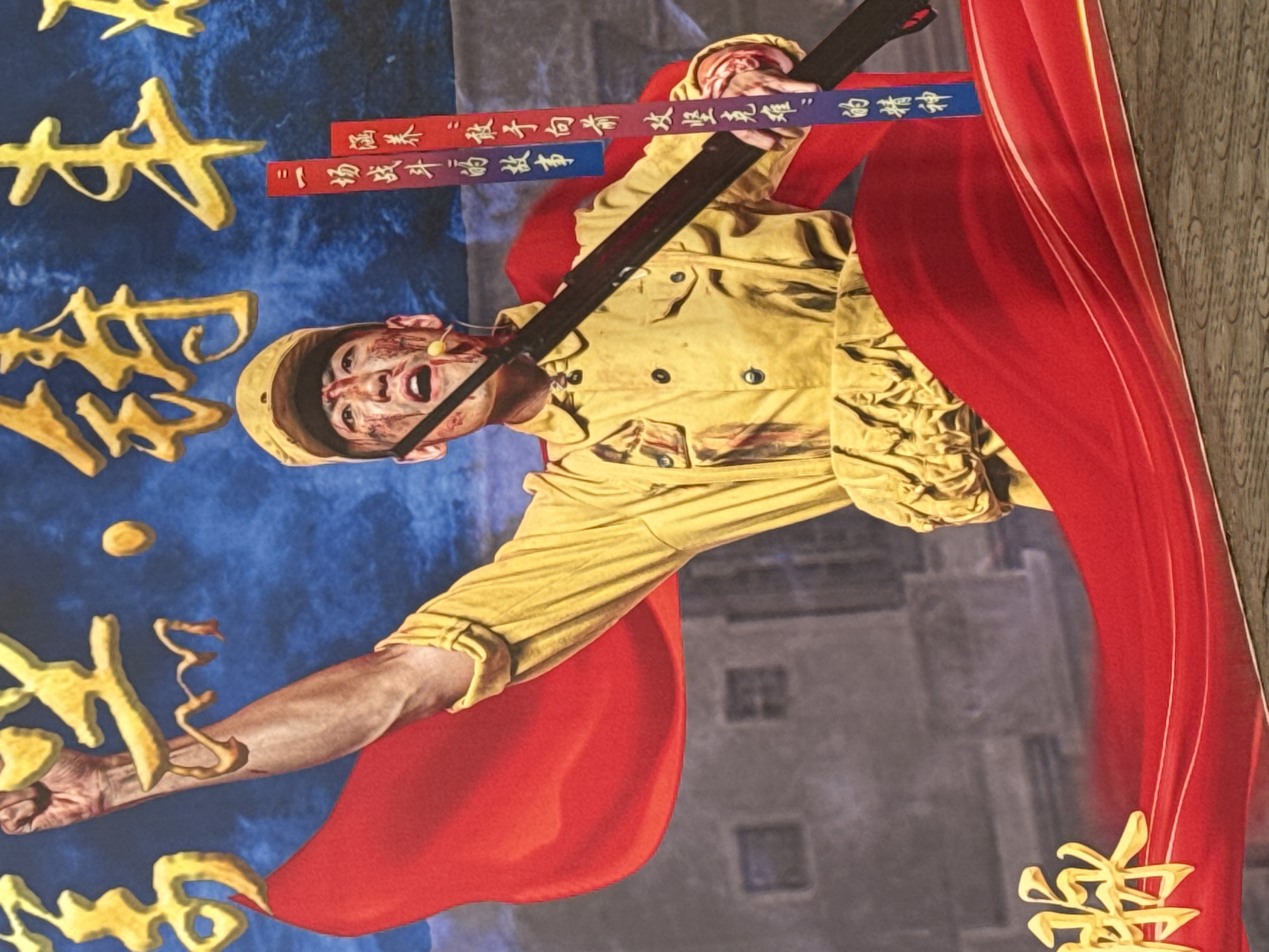 This vibrant and dramatic artwork features a male figure, likely a soldier or revolutionary, in a dynamic, action-oriented pose. He wears a yellow military-style jacket and cap, and his face is streaked with blood, suggesting injury or intense struggle. His mouth is open, possibly in a shout or cry, and he has a small, light-colored object, perhaps a bullet or a prop, lodged in his mouth. He is holding a long, dark rifle or sword with both hands, positioned across his body.
A large, flowing red fabric, resembling a cape or banner, dramatically sweeps around him, contrasting with the deep blue background on the left, which is adorned with stylized golden Chinese characters. On the right, faint grey rectangular shapes in the background suggest buildings or structures.
Two ribbon-like banners are held by the figure, one red and one blue. The red banner displays Chinese text: "涵养“敢于向前攻坚克难”的精神" (Cultivate the spirit of "daring to move forward, overcome difficulties and challenges"). The blue banner has partially visible characters that likely continue or relate to this theme. On the blue background, golden characters are partially visible, including "一场战斗的故事" (A story of a battle) and "脉".
The overall scene conveys intense emotion, heroism, and determination in the face of adversity, characteristic of propaganda art or a theatrical poster. The man's expression and the weapon suggest a moment of fierce combat or sacrifice.