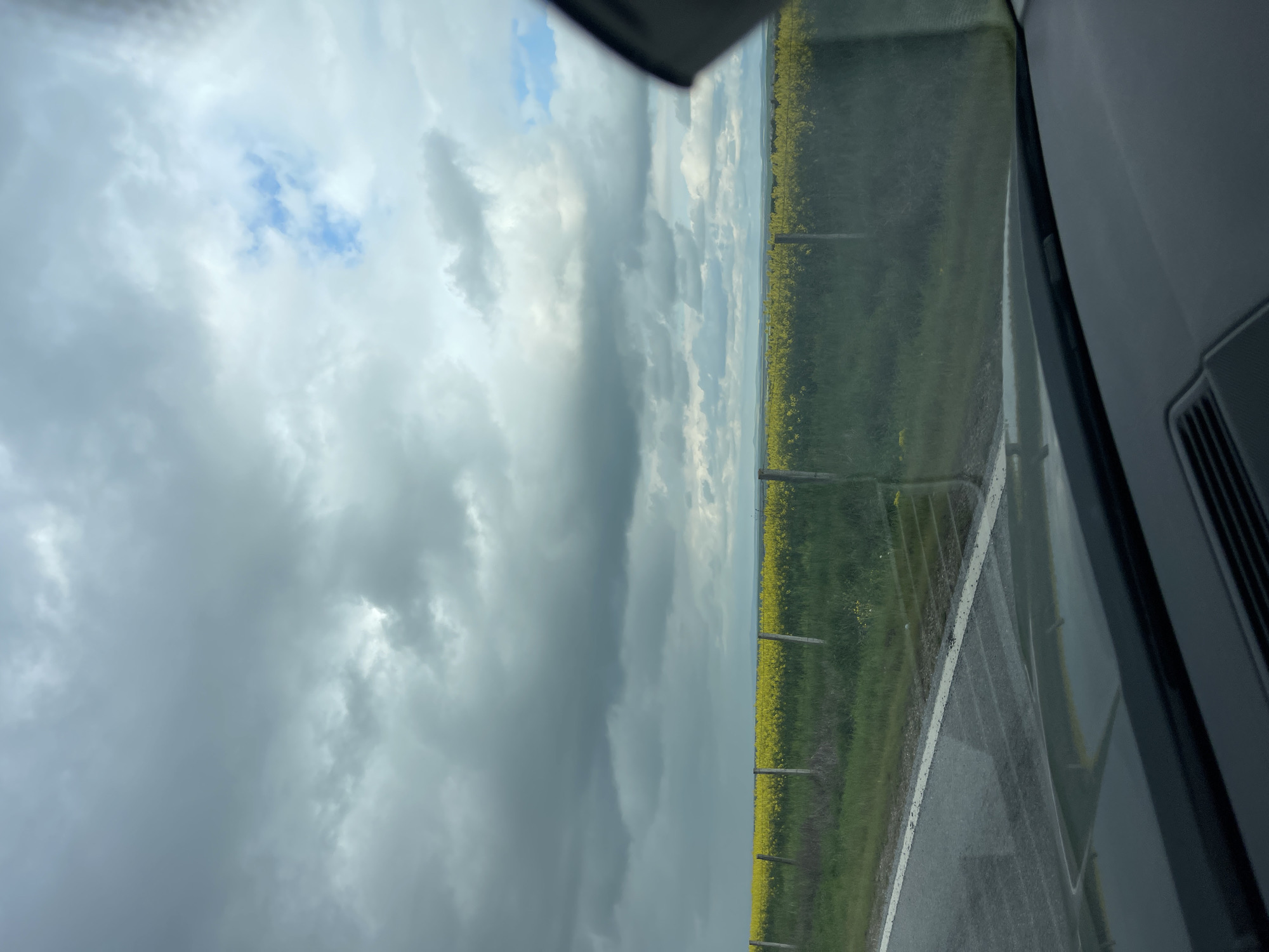 This landscape photograph captures a rural scene from the perspective of a vehicle, possibly on a road trip near Vodenichane, Bulgaria. The image is composed of three main horizontal sections: a road in the foreground, vast yellow fields in the midground, and a dramatic sky above.
On the right side of the frame, elements of a car's interior are visible, including a portion of the dark dashboard, a side pillar, and a vent, indicating the vantage point is from within a vehicle. A paved road, appearing slightly damp or wet, runs along the bottom and right edges, marked by a clear white solid line.
Separating the road from the agricultural land is a simple fence, with several wooden posts visible. Beyond this, a wide field of bright yellow flowering crops, characteristic of canola or rapeseed, stretches horizontally across the image, extending towards the distant horizon. The vibrant yellow contrasts with patches of green vegetation.
The sky occupies the upper portion of the photograph, dominated by large, impressive grey and white clouds that suggest an overcast or partly cloudy day. Breaks in the cloud cover reveal patches of clearer blue sky, particularly towards the upper left. A subtle line of distant hills or land is visible where the fields meet the sky on the horizon.
No people or visible text are present in the image. The time of day appears to be daytime, with a soft, diffused light due to the cloud cover. The overall impression is one of expansive agricultural land under a dynamic sky, viewed during travel through a rural region.