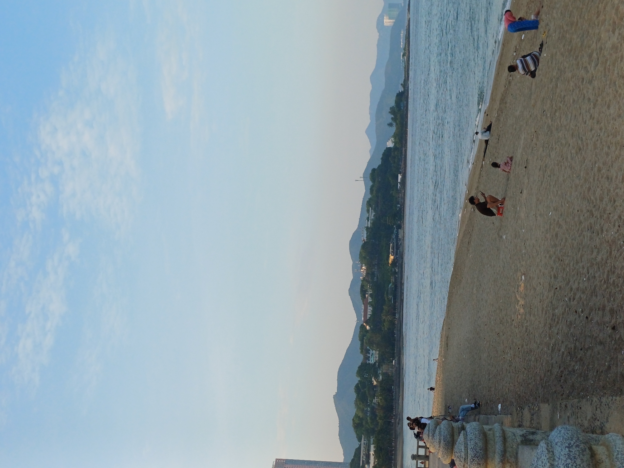 A wide shot captures a sandy beach in Nha Trang, Vietnam, with a calm sea lapping at the shore. The sky is a soft blue with scattered white clouds. In the background, rolling green hills are dotted with houses and trees, and distant mountains fade into the horizon. A few modern buildings, including a tall, slender skyscraper, are visible on the coastline. Several people are scattered across the beach. On the left, a group of individuals stands near a stone barrier, with some sitting on it. Further down the beach, a person kneels, appearing to use a mobile phone. Nearby, a child in a striped outfit sits on the sand. Another person sits with their back to the viewer, facing the sea, while a blue-clad figure bends over in the foreground.