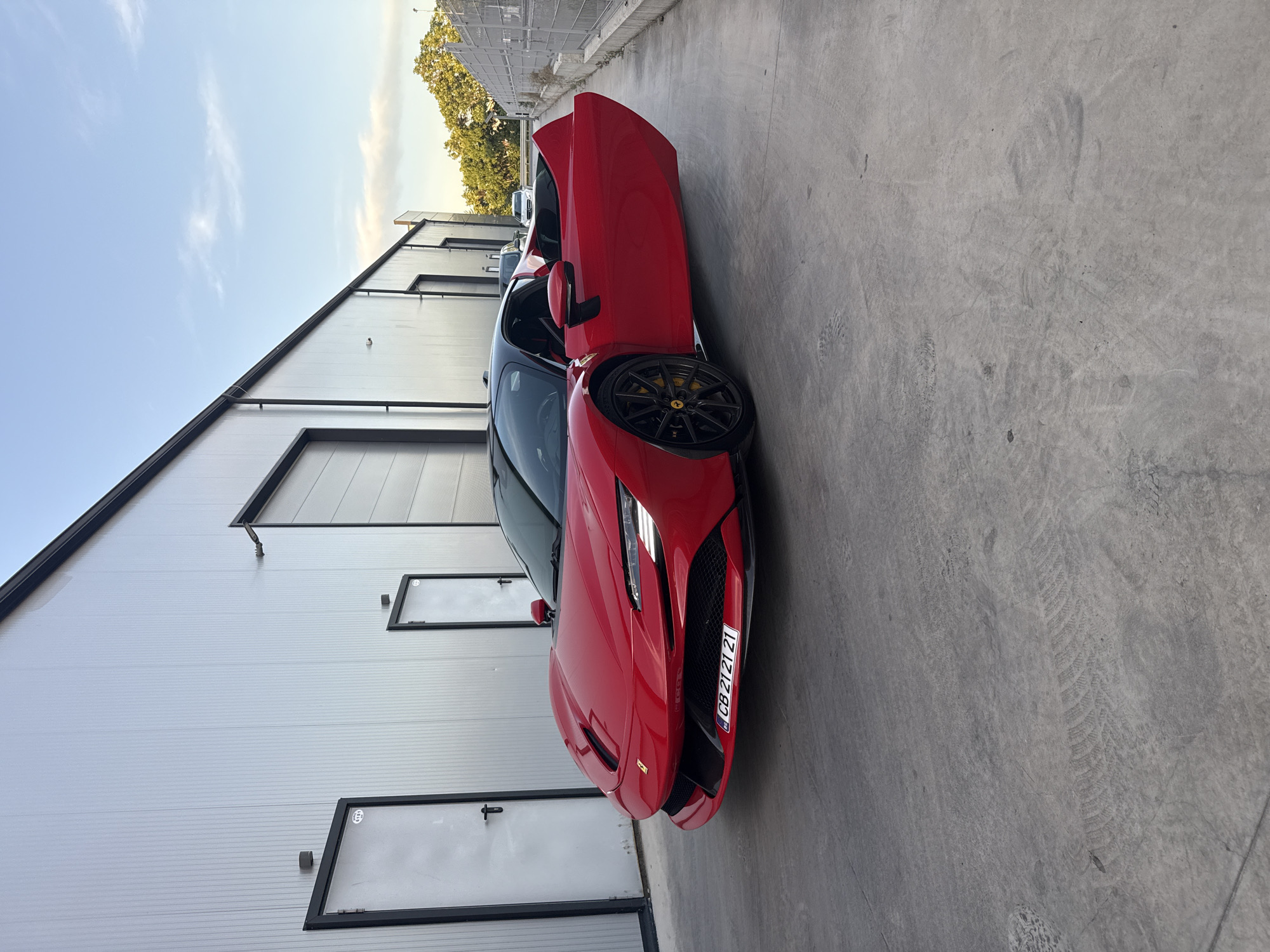 A red Ferrari is parked on a concrete surface in front of a light gray building with multiple metal doors and small rectangular windows with black frames. The driver-side door of the car is open. The car's license plate is visible. The scene appears to be in an industrial area. A small patch of trees can be seen in the background. The image was captured on a clear day with a blue sky. Due to the license plate, it can be identified to be in Plovdiv, Bulgaria.