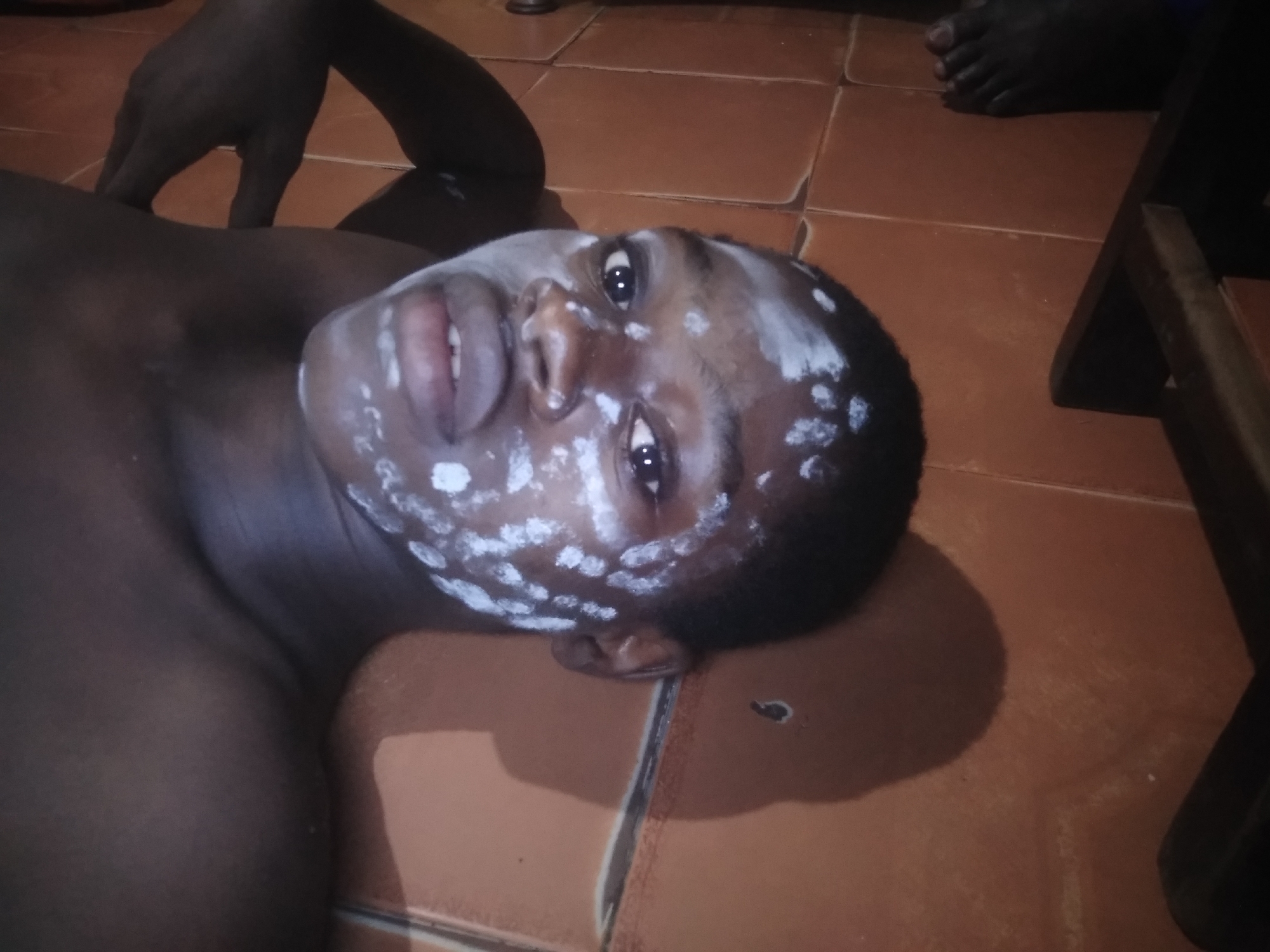 A dark-skinned person lies on a tiled floor. The person's face is decorated with white spots, and their eyes are open. The person is partially undressed. The floor is covered in brown square tiles. A dark-skinned foot is visible in the upper right corner. A part of a wooden chair sits in the lower right corner. The location is potentially a private residence in Shangisha, Nigeria.