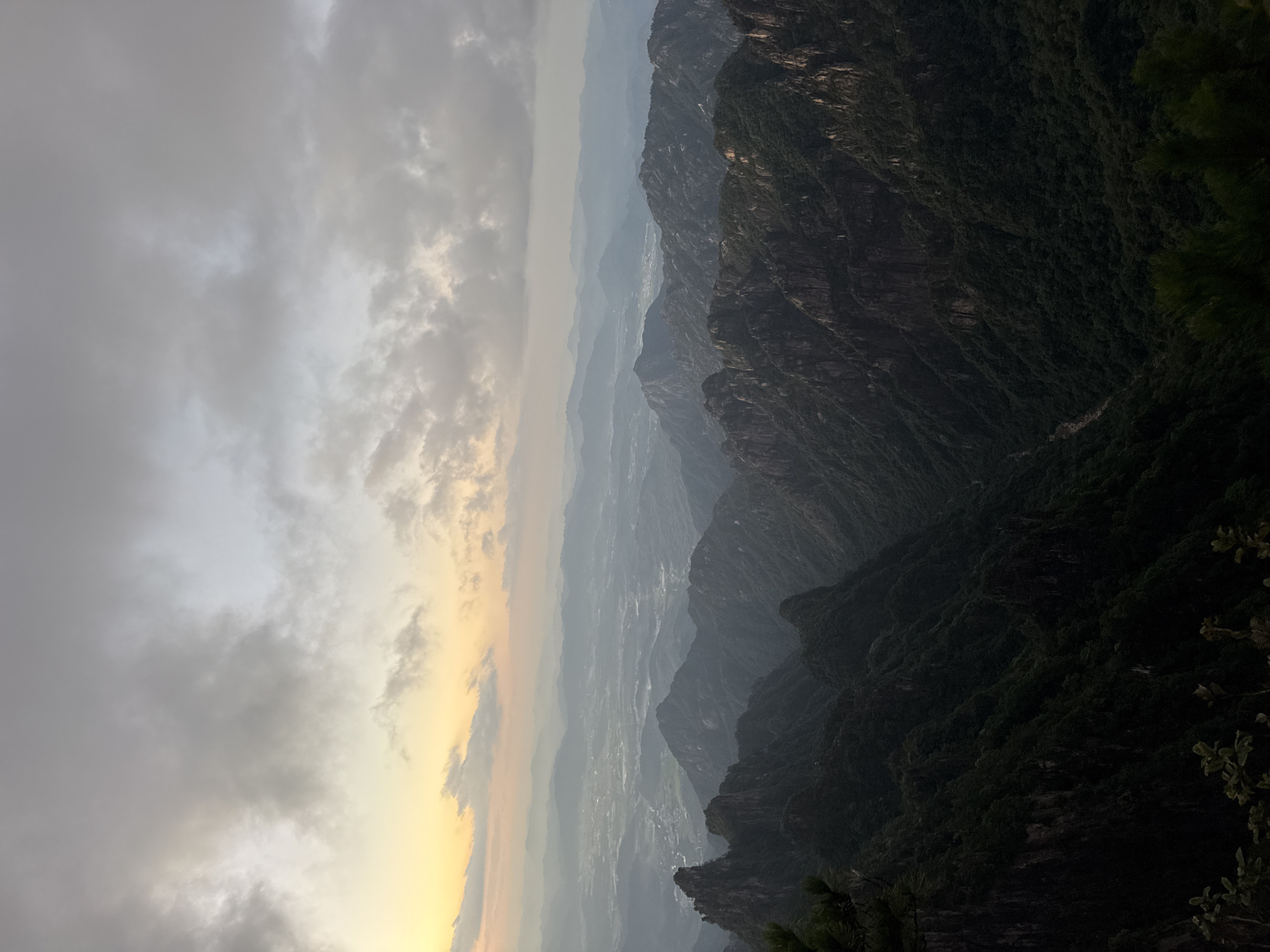 The image captures a panoramic view of a mountainous landscape in the city of Huangshan District, China, likely during sunrise or sunset. The foreground features dark, rugged mountain peaks densely covered in lush green vegetation. The midground reveals more layers of mountains receding into the distance, their forms softened by atmospheric haze. The background shows a valley with hints of human settlements, possibly a town or village, nestled amongst the undulating terrain. The sky above is filled with a gradient of colors, from a soft yellow and orange near the horizon, transitioning to a pale grey and white with scattered clouds higher up. The overall impression is one of natural grandeur and the serene beauty of a mountain vista at dusk or dawn. There are no people or specific objects clearly identifiable in the scene, focusing solely on the vast natural environment.