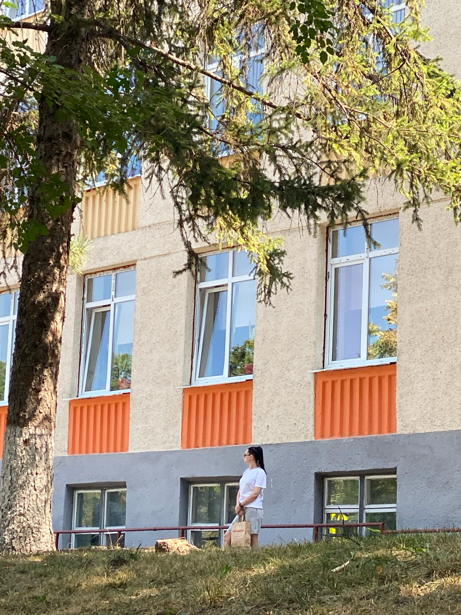 A young woman wearing a white t-shirt and light-colored shorts stands in front of a building in Chișinău, Moldova. She is looking to her left, with a paper shopping bag in her right hand. The building has several large windows and is painted in shades of beige and orange. A large tree with green foliage is in the foreground, partially obscuring the view of the building. The ground in front of the building is covered in grass and some debris. The overall lighting suggests it is daytime, with the sun casting shadows.