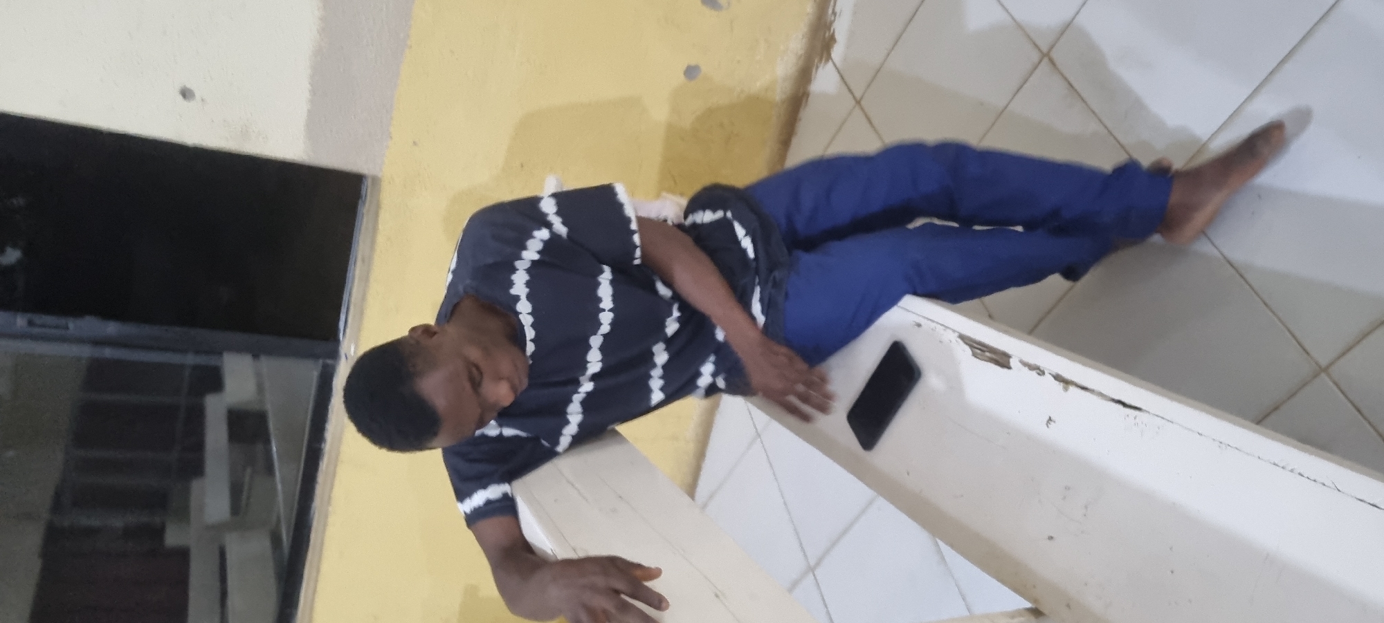 A Black male, appearing to be in his late teens or early twenties, is seated on a white, slatted bench in an indoor location. He is wearing a dark t-shirt with horizontal white stripes and blue trousers. His head is bowed, and he is looking down. His right arm rests on the back of the bench. A black smartphone is placed on the bench in front of him. The background consists of a yellow wall with some circular marks and a dark, reflective surface that appears to be a window or door. The floor is tiled in a diamond pattern. The overall lighting suggests it is indoors, possibly at night or in a dimly lit area. The scene does not provide specific visual cues to pinpoint a location within Lagos, Nigeria, beyond it being an urban indoor setting.